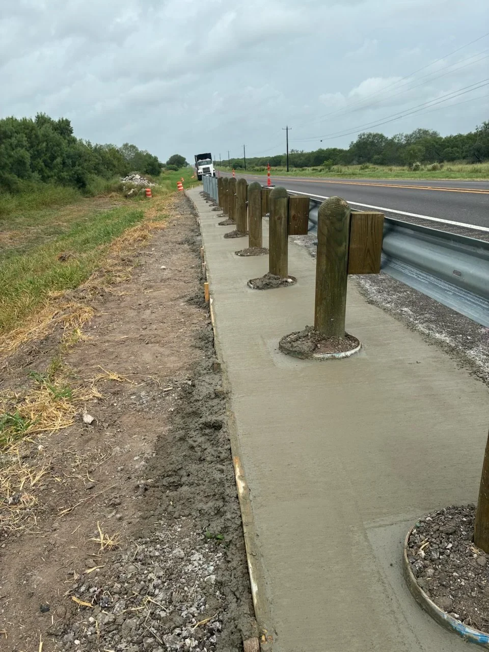 Construction site where a sidewalk is being built along a highway, with wooden posts set in concrete along the edge, and a concrete curb in place.