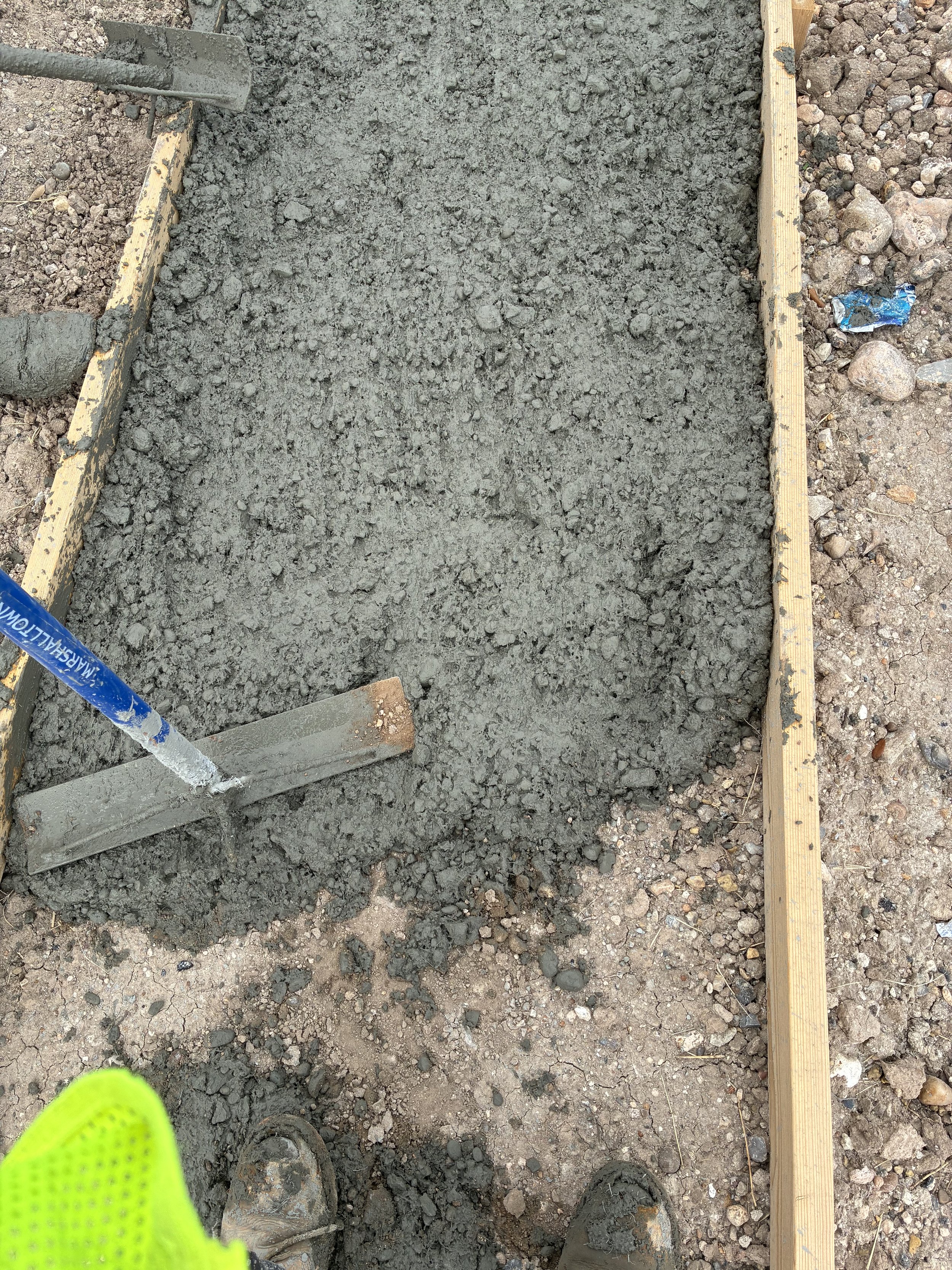 A construction site with freshly poured concrete within wooden forms, surrounded by dirt and gravel, with construction tools and materials visible.