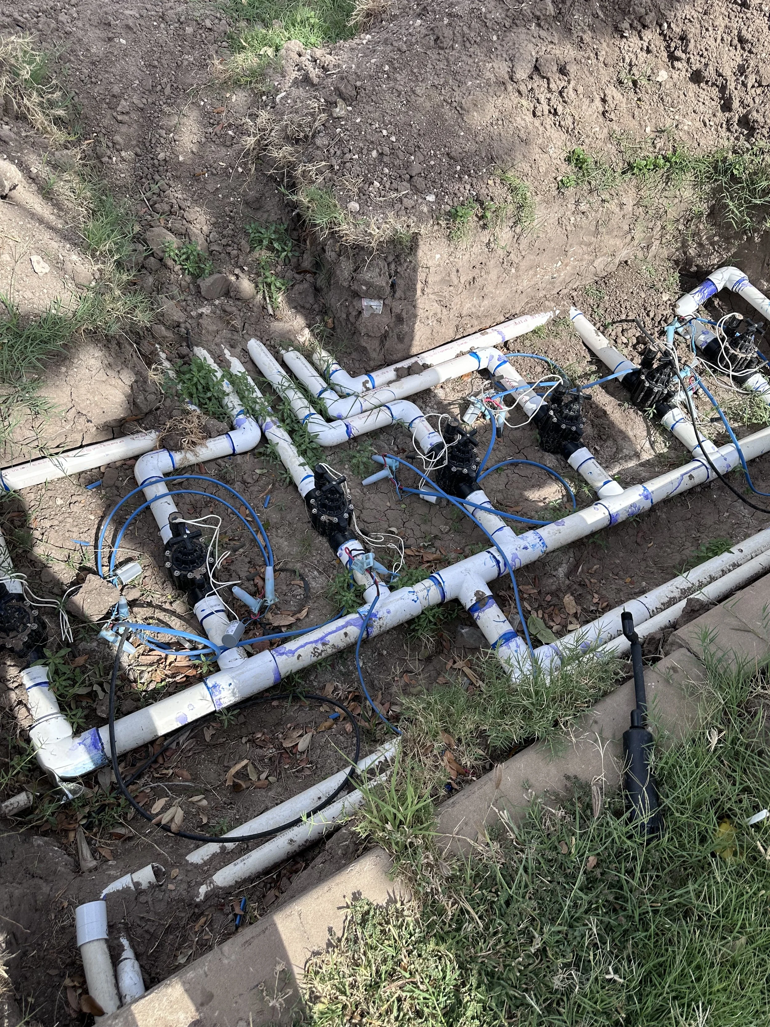 A broken underground sprinkler system with white PVC pipes, valves, and blue wiring, situated in a patch of dirt and grass.