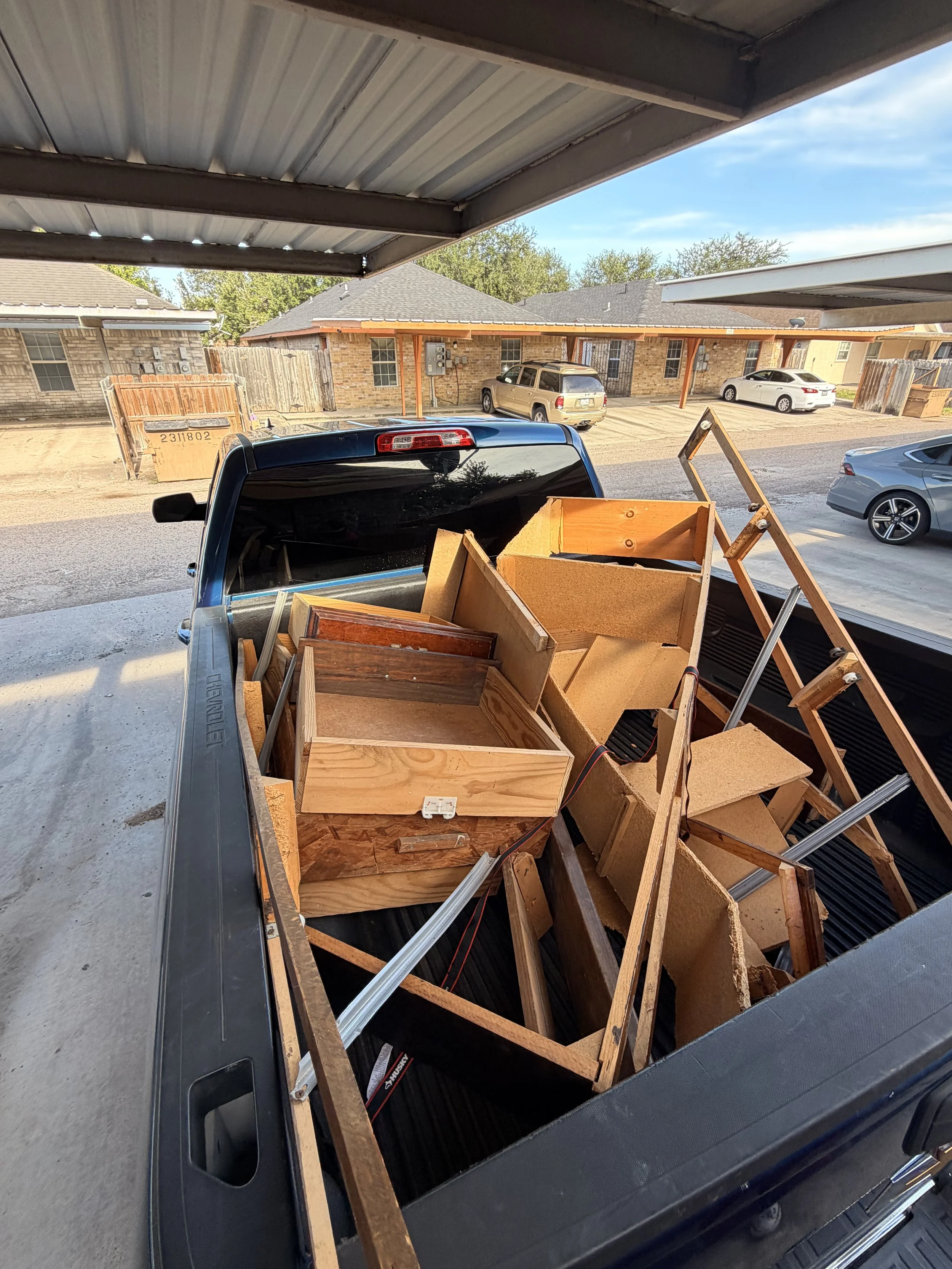 The image shows the bed of a pickup truck filled with wooden furniture pieces, including cabinets and shelves, parked in a residential driveway with a covered porch and neighboring houses in the background.