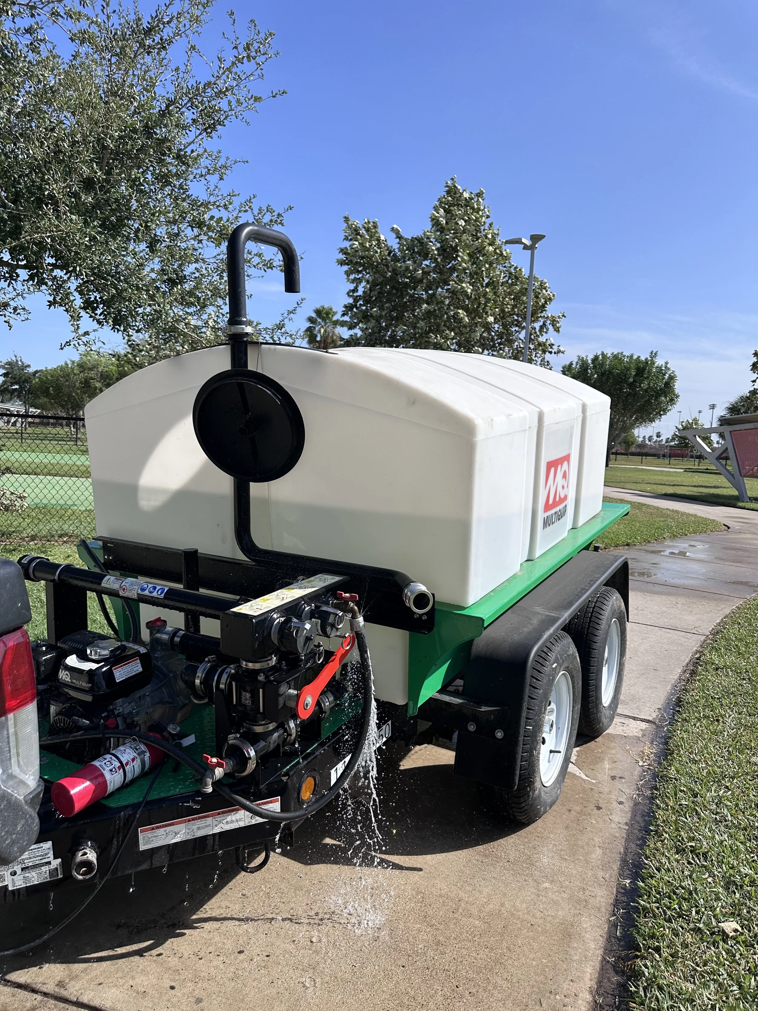 A utility cart with a large white water tank and firefighting equipment, including fire extinguishers and hoses, parked outdoors on a paved area with grass, trees, and a blue sky in the background.