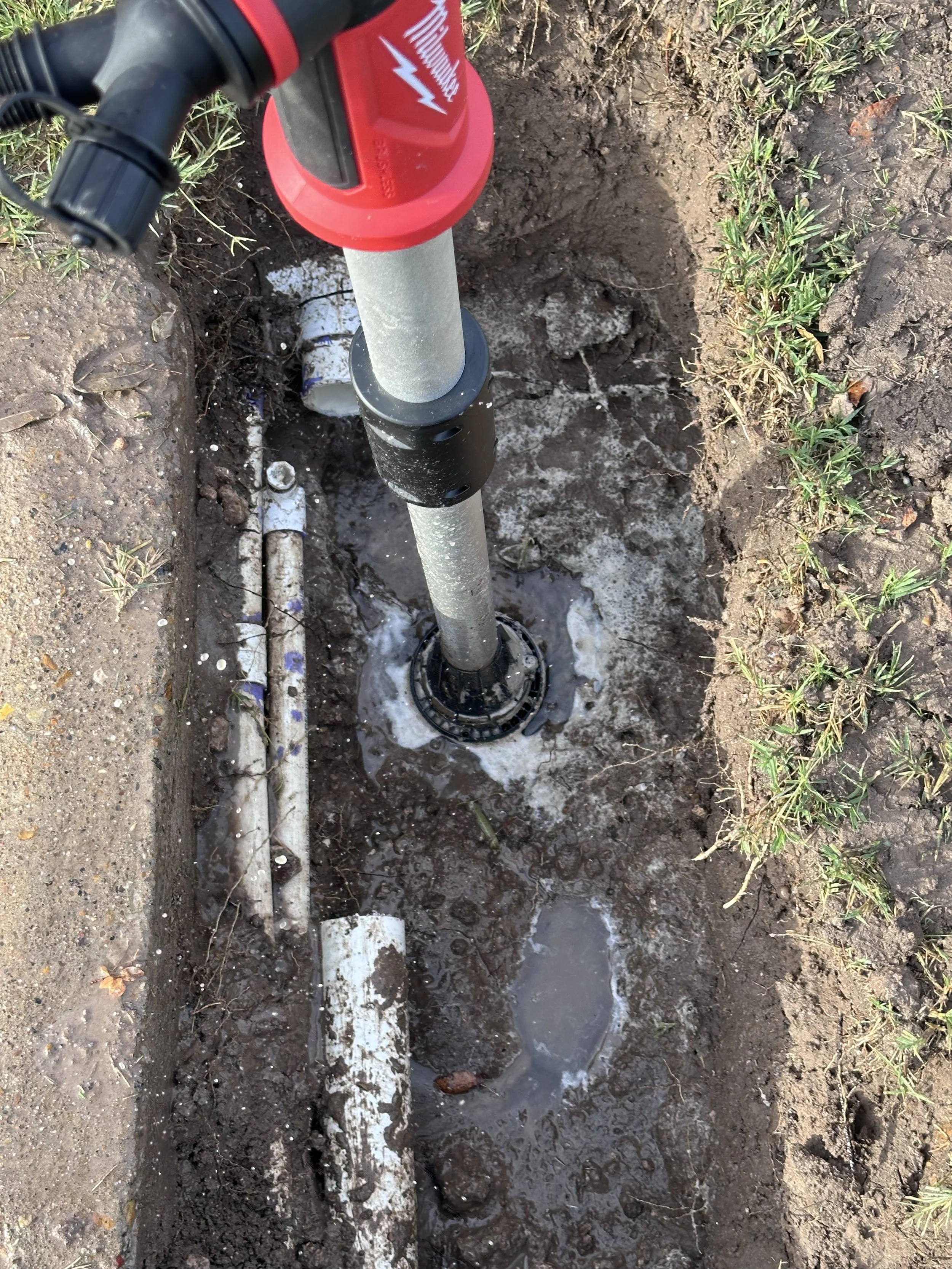 A close-up view of a buried utility or water pipe installation in an open trench with soil and grass around.