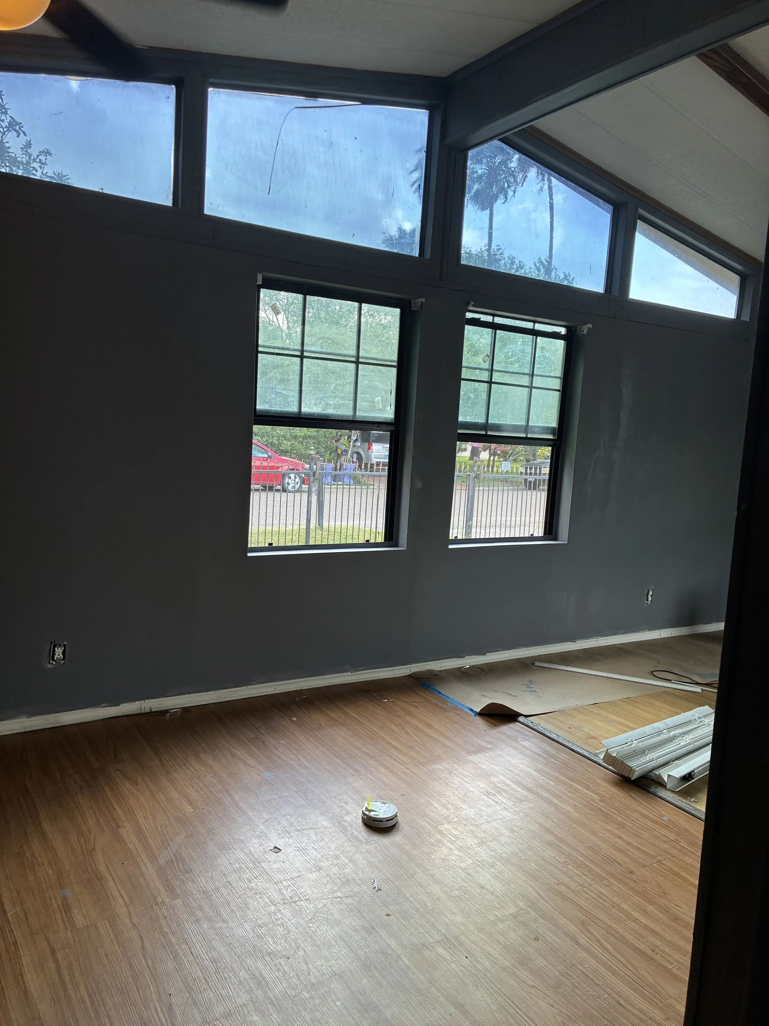 An interior room with a hardwood floor, gray walls, and large windows that let in natural light. Some construction or renovation materials are on the floor, and a small round device, possibly a robot vacuum or sensor, is near the center.