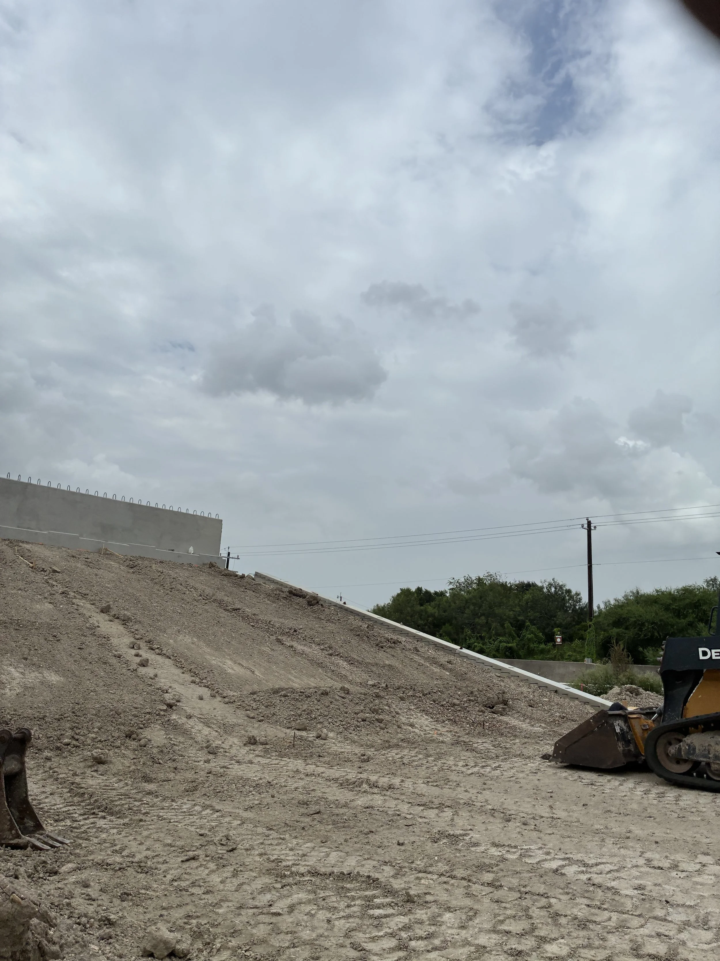 Construction site with dirt ground, partially built wall, and construction equipment, cloudy sky overhead, power lines, trees in the background.