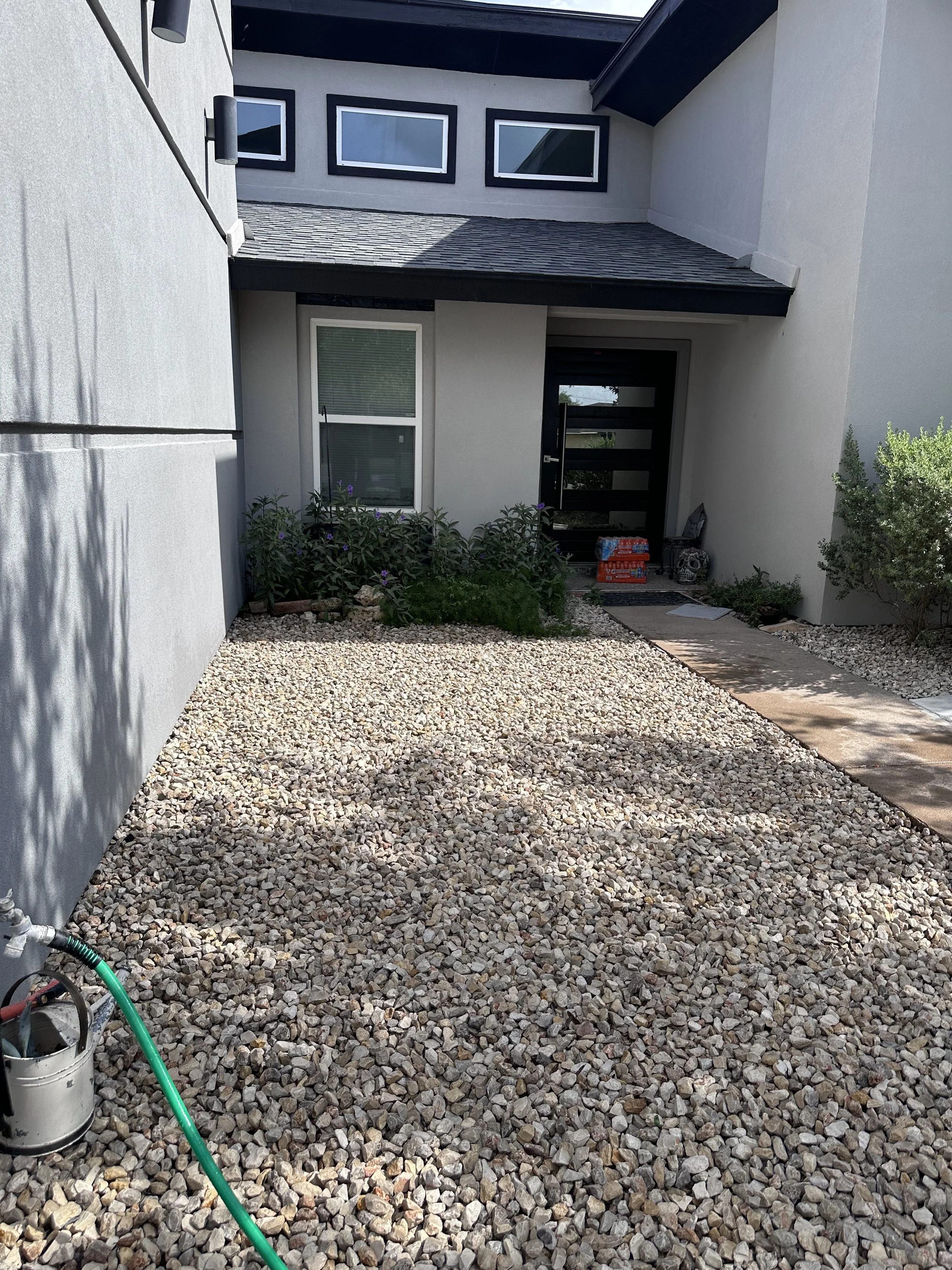 Front yard gravel garden with a small pathway, plants, and a house entrance with a black door and window blinds.