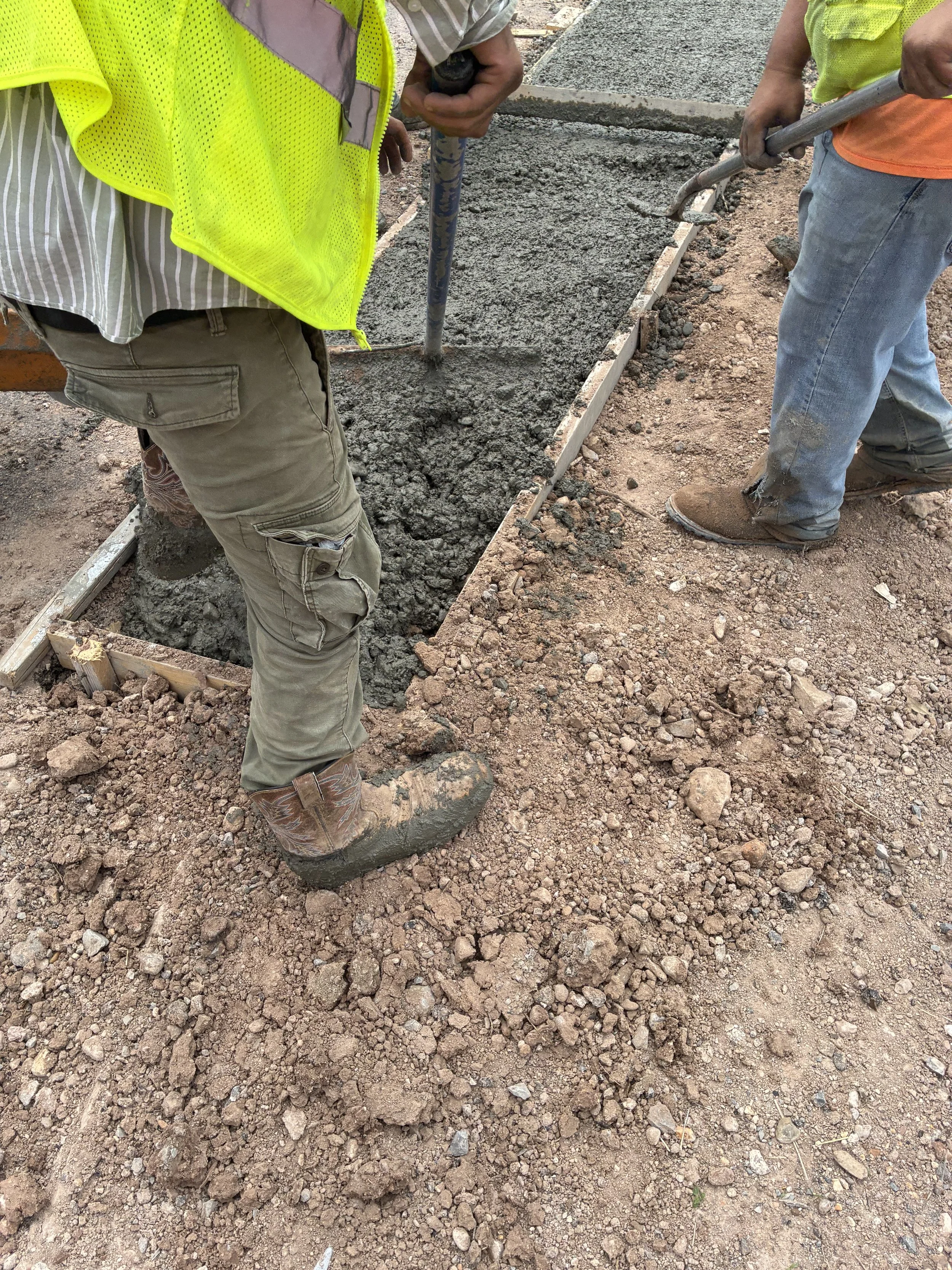 Two construction workers are working on pouring concrete for a sidewalk. One worker is wearing cargo pants, a striped shirt, a bright yellow safety vest, and cowboy boots. The other worker is wearing blue jeans, an orange shirt, a bright yellow safet