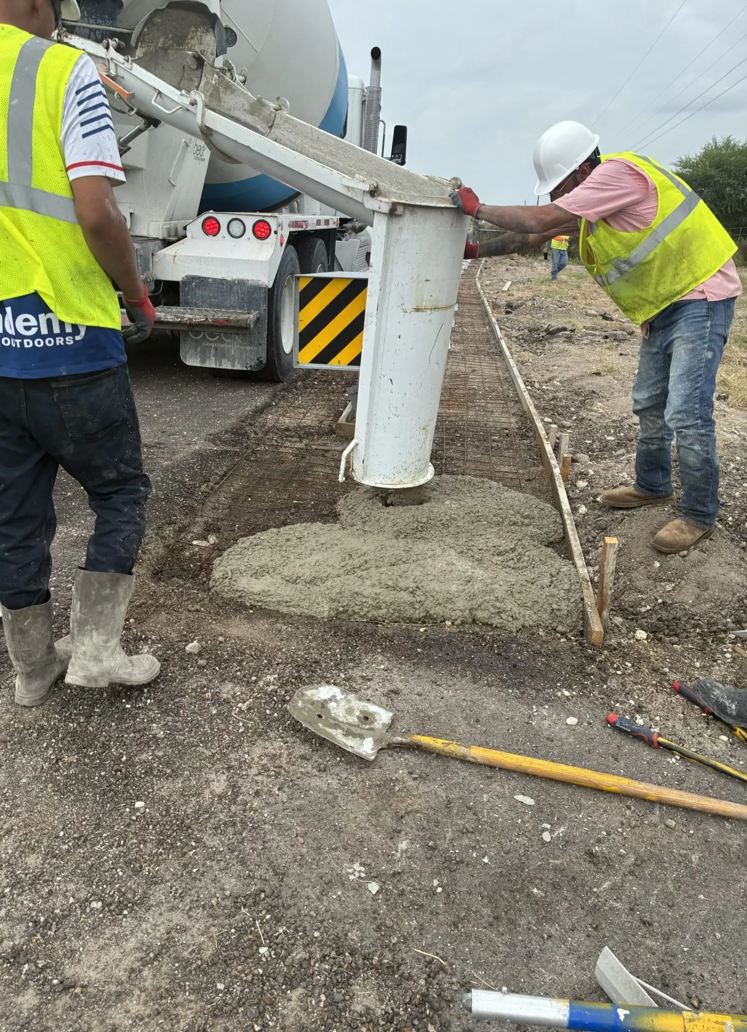 Construction workers in safety vests and helmets pouring concrete at a construction site, with tools and dirt on the ground.