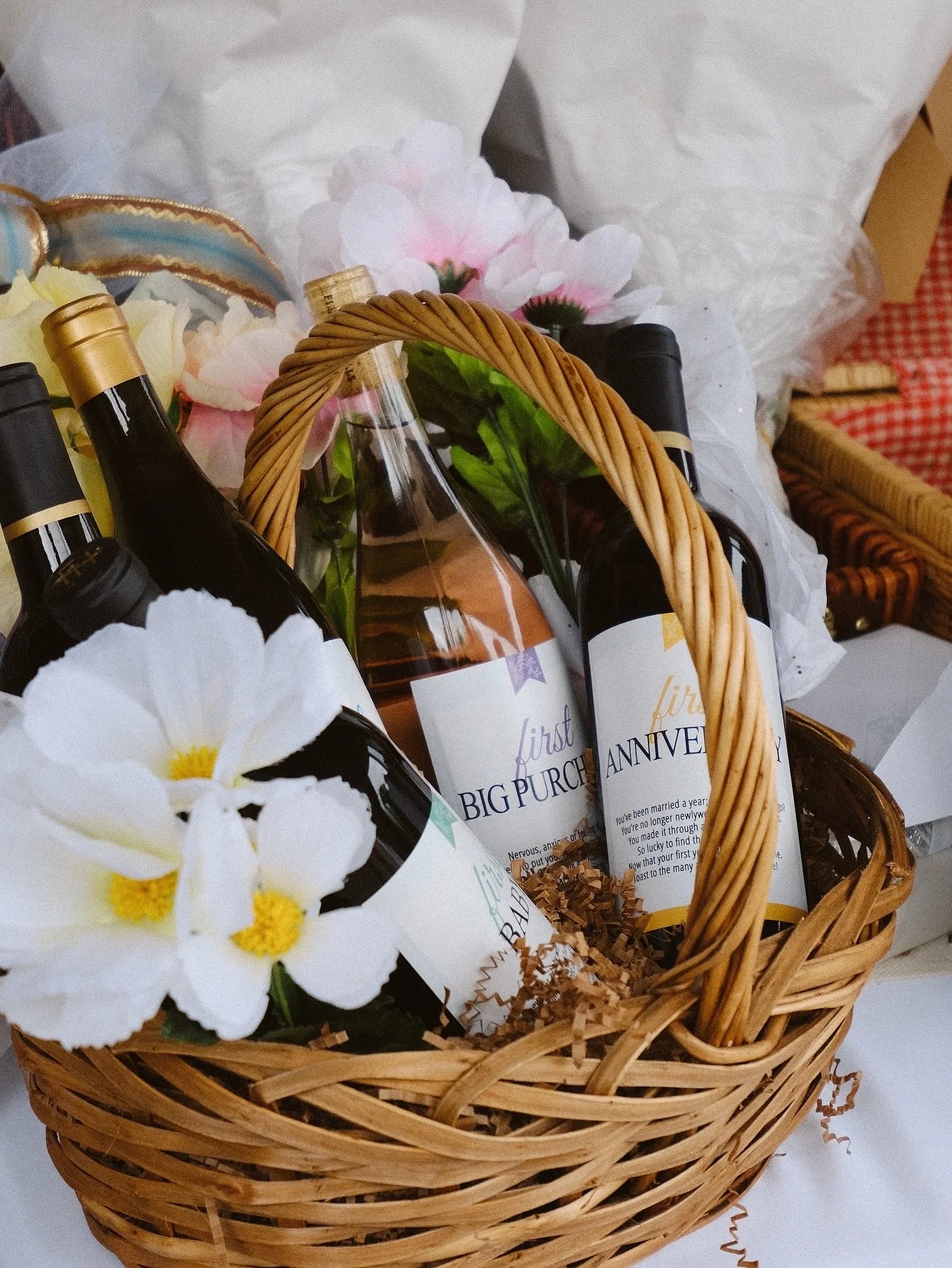 A wicker basket filled with bottles of wine, pink and white flowers, and printed cards celebrating a first anniversary, surrounded by tissue paper and decorative paper shreds.