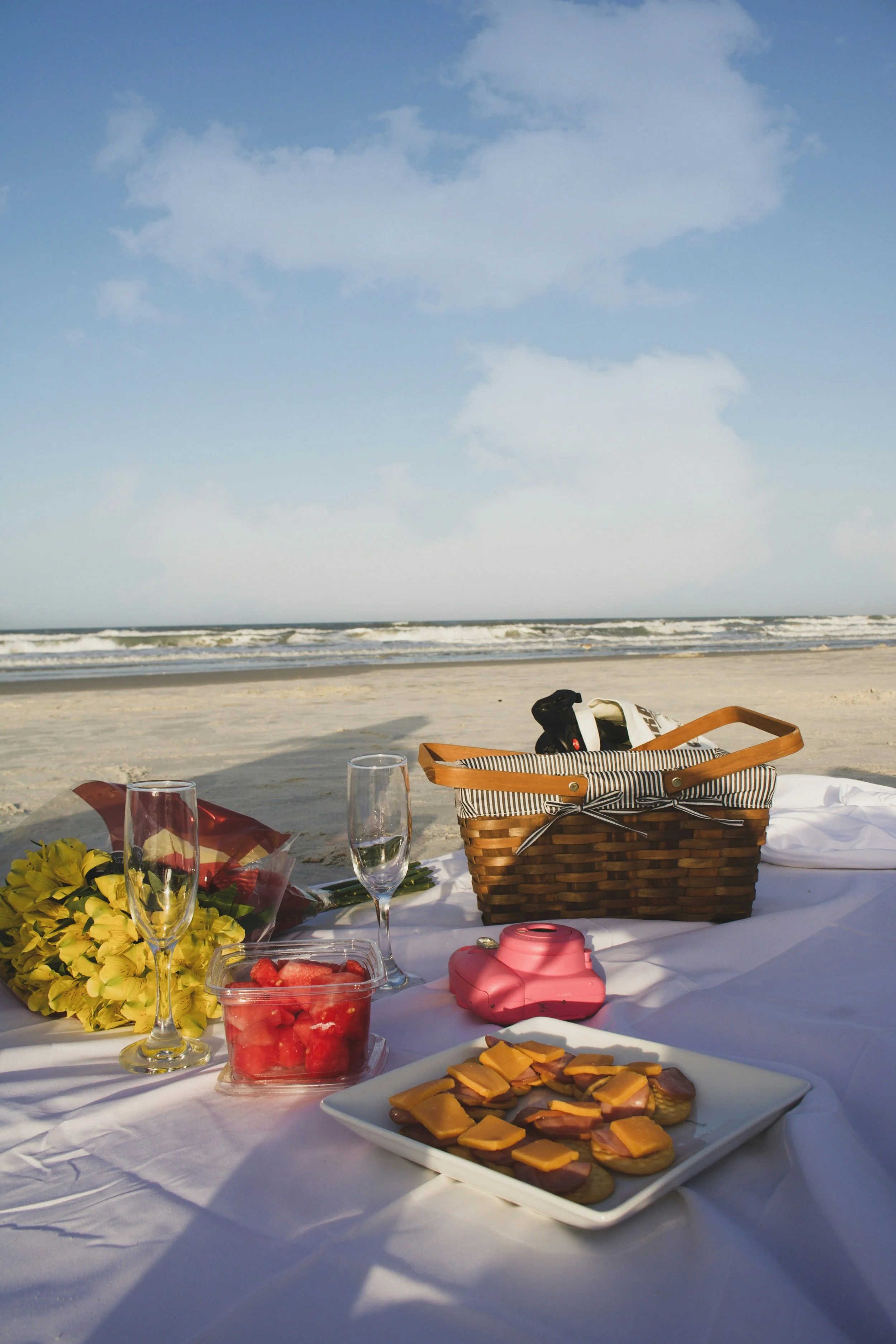 Picnic table on the beach with food and drinks, including cheese and crackers, watermelon, and flowers, with the ocean and sky in the background.