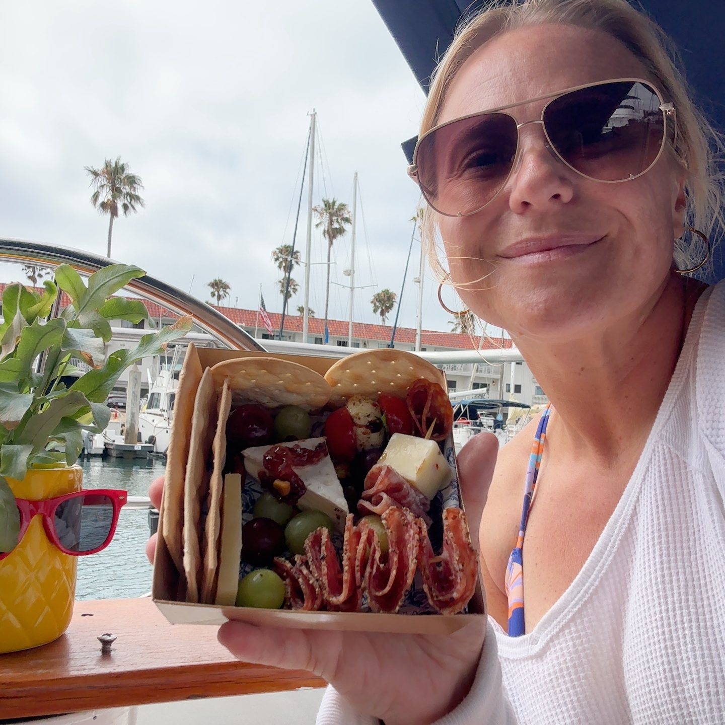 Woman smiling, wearing sunglasses, on sailboat, holding a box of assorted snacks and cheeses, with Oceanside marina and palm trees in the background.