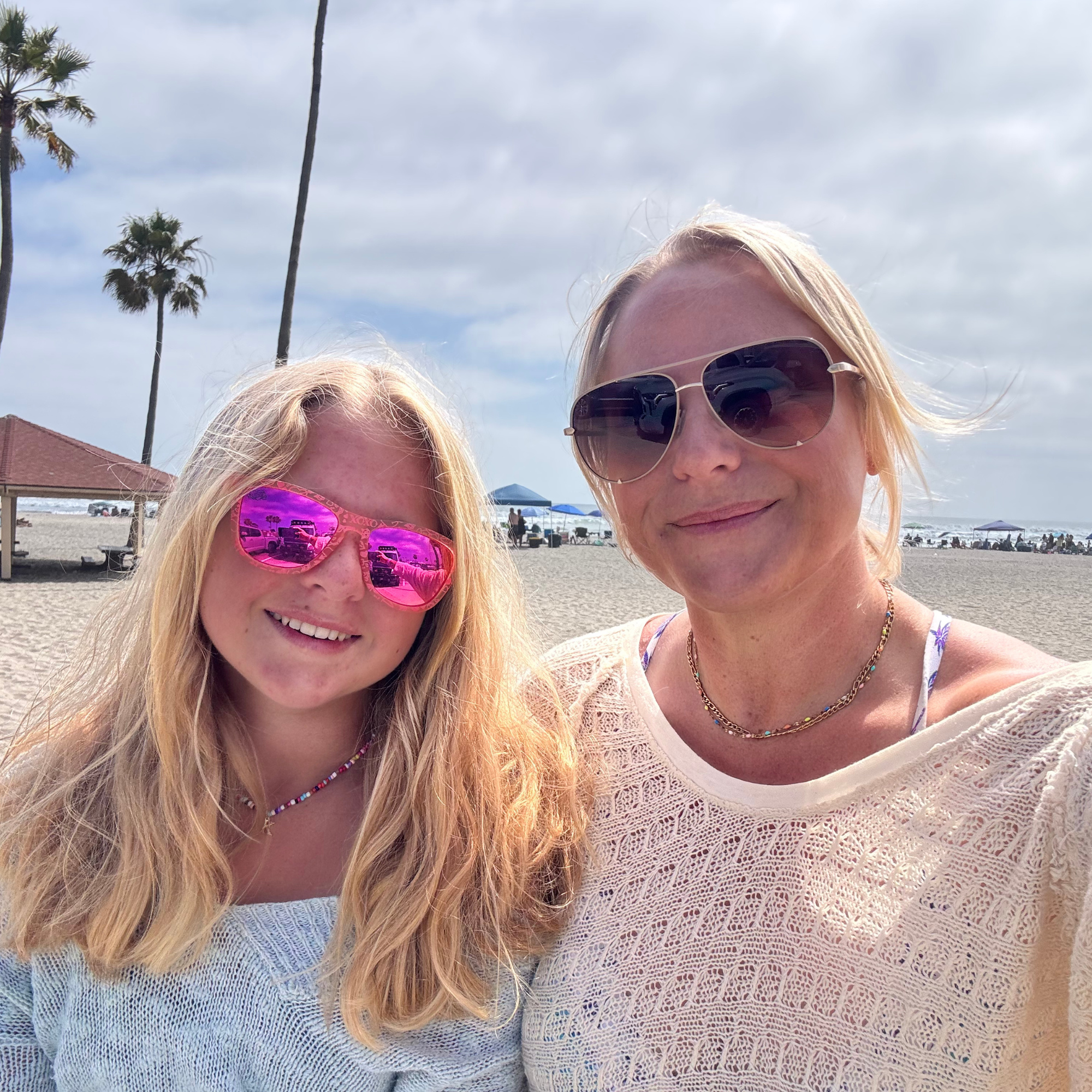 Two women taking a selfie at the beach, wearing sunglasses and smiling, with palm trees and umbrellas in the background under a cloudy sky.