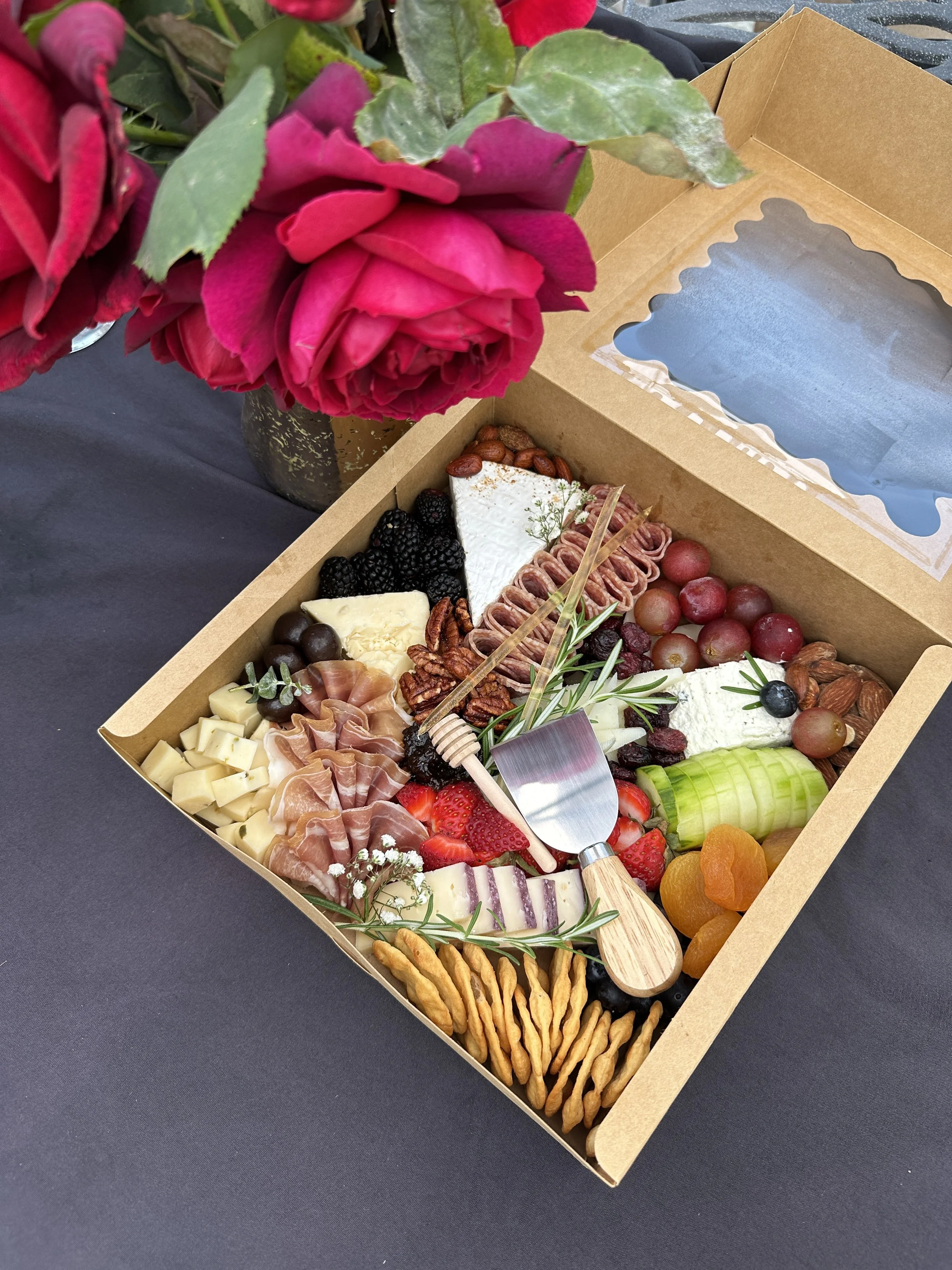 A box containing a variety of cheeses, grapes, nuts, and meats, with a glass of water inside the lid.