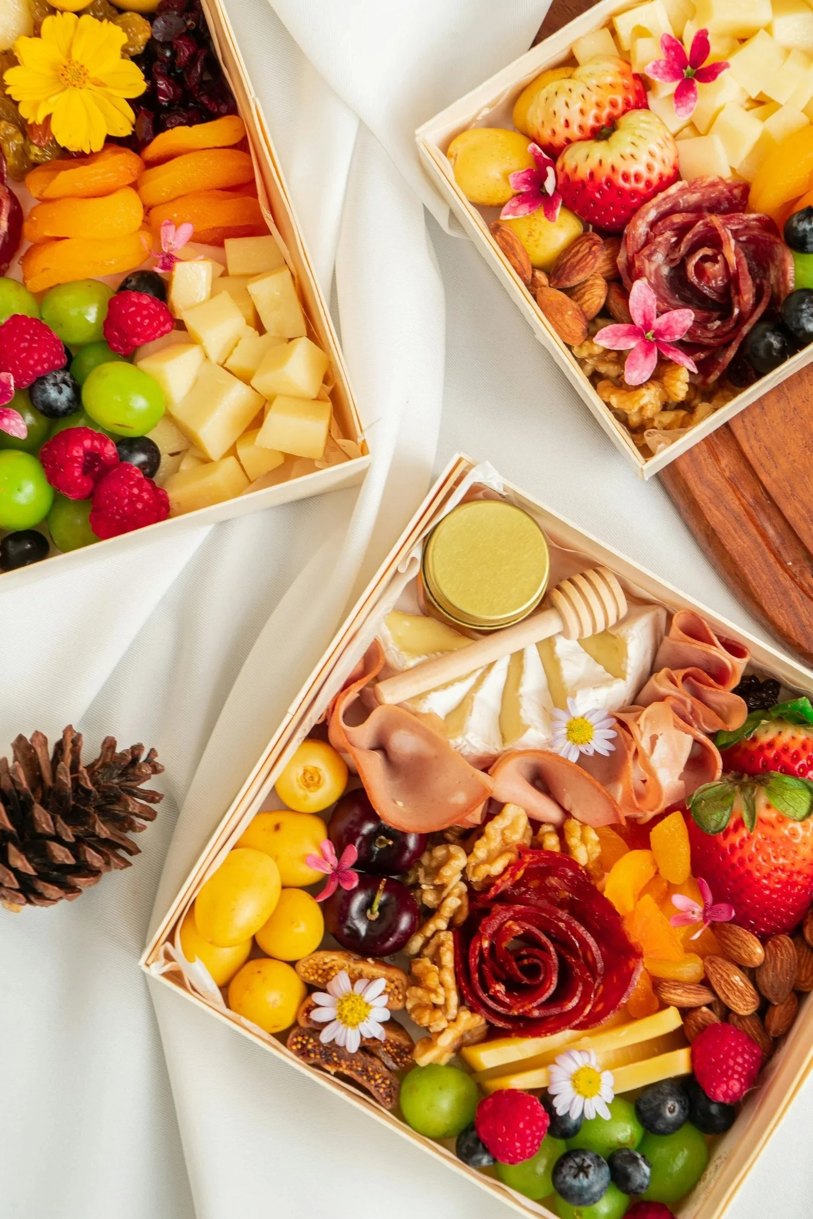 A gift box filled with an assortment of fresh fruit, nuts, cheese, and edible flowers, arranged as a gourmet snack or gift.
