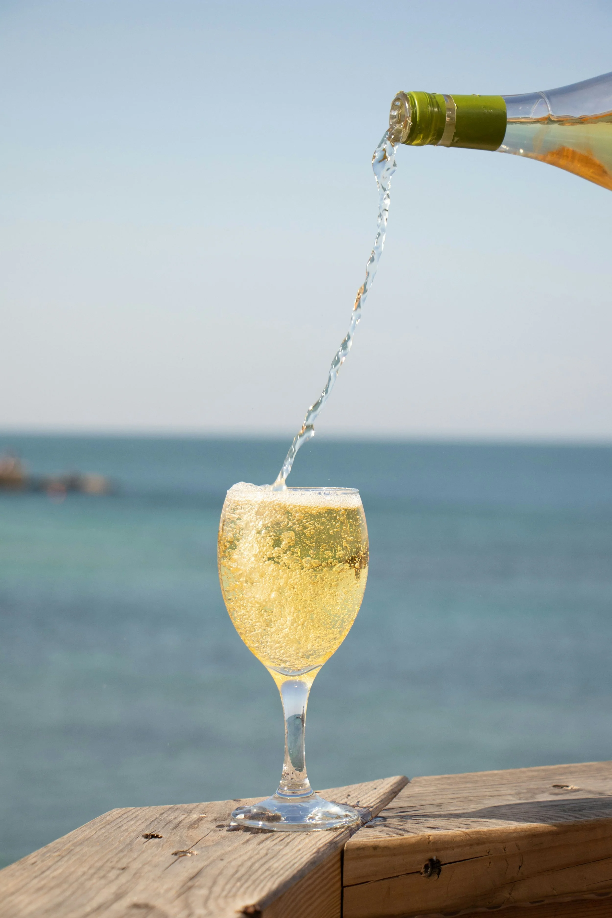 A glass of sparkling champagne on a wooden surface with a bottle pouring champagne into it, with an ocean and clear sky in the background.