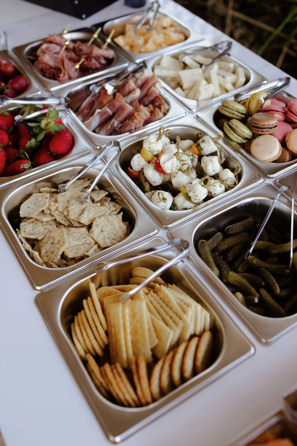 A bottomless charcuterie buffet cart with various snacks including strawberries, crackers, mozzarella balls, pickles, cookies, cheese, cured meats, and fruit salad in metal containers.