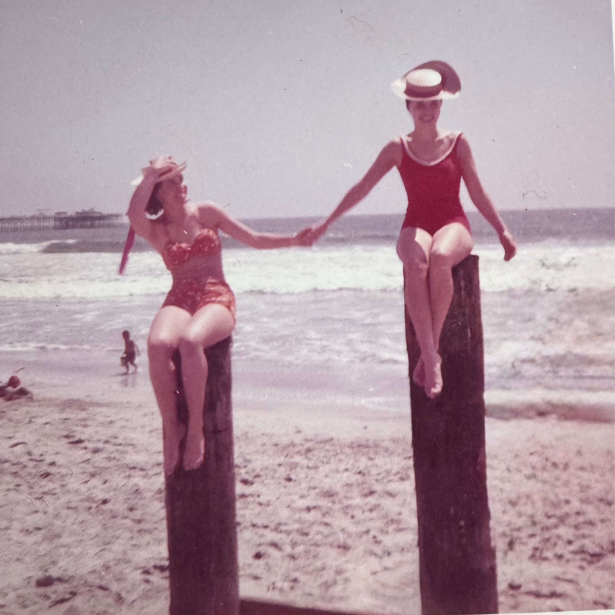 A vintage color photograph of two women in swimsuits holding hands on a beach, with the ocean and a pier in the background, and a hat on one woman. Girl on right is Annette La Mor Eastman.