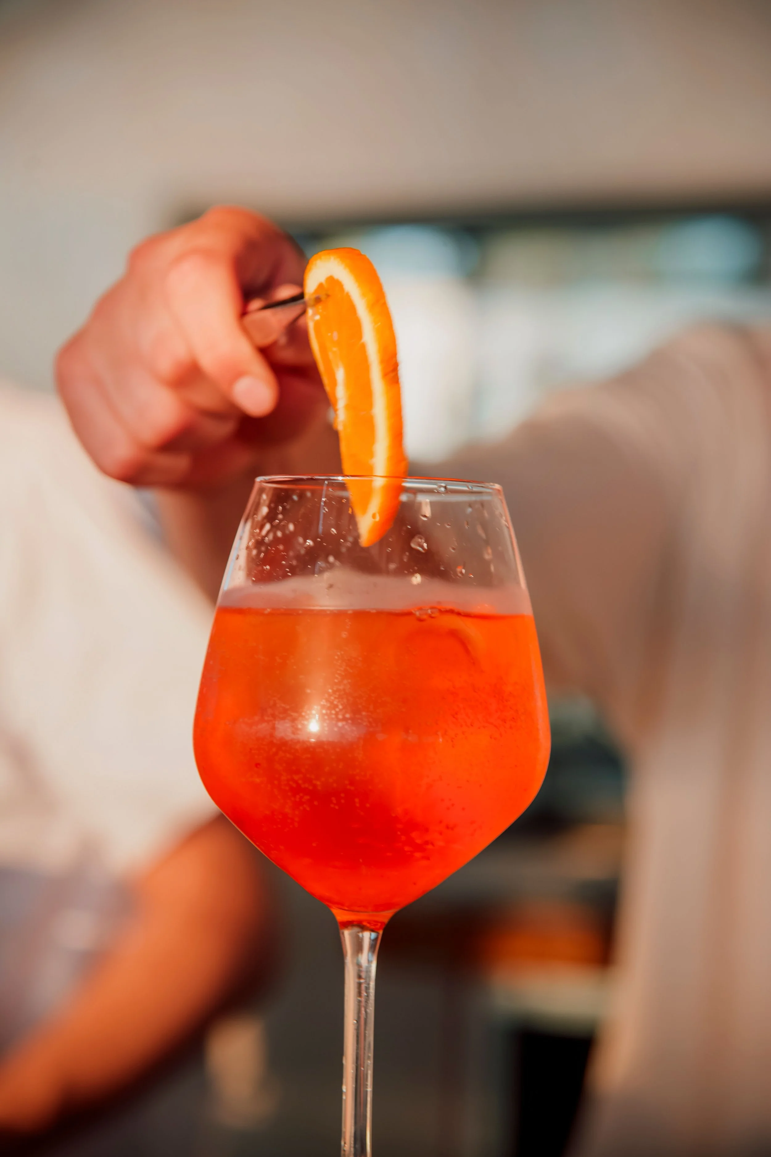 A hand garnishing an orange-colored cocktail with a slice of orange on the rim of a glass.