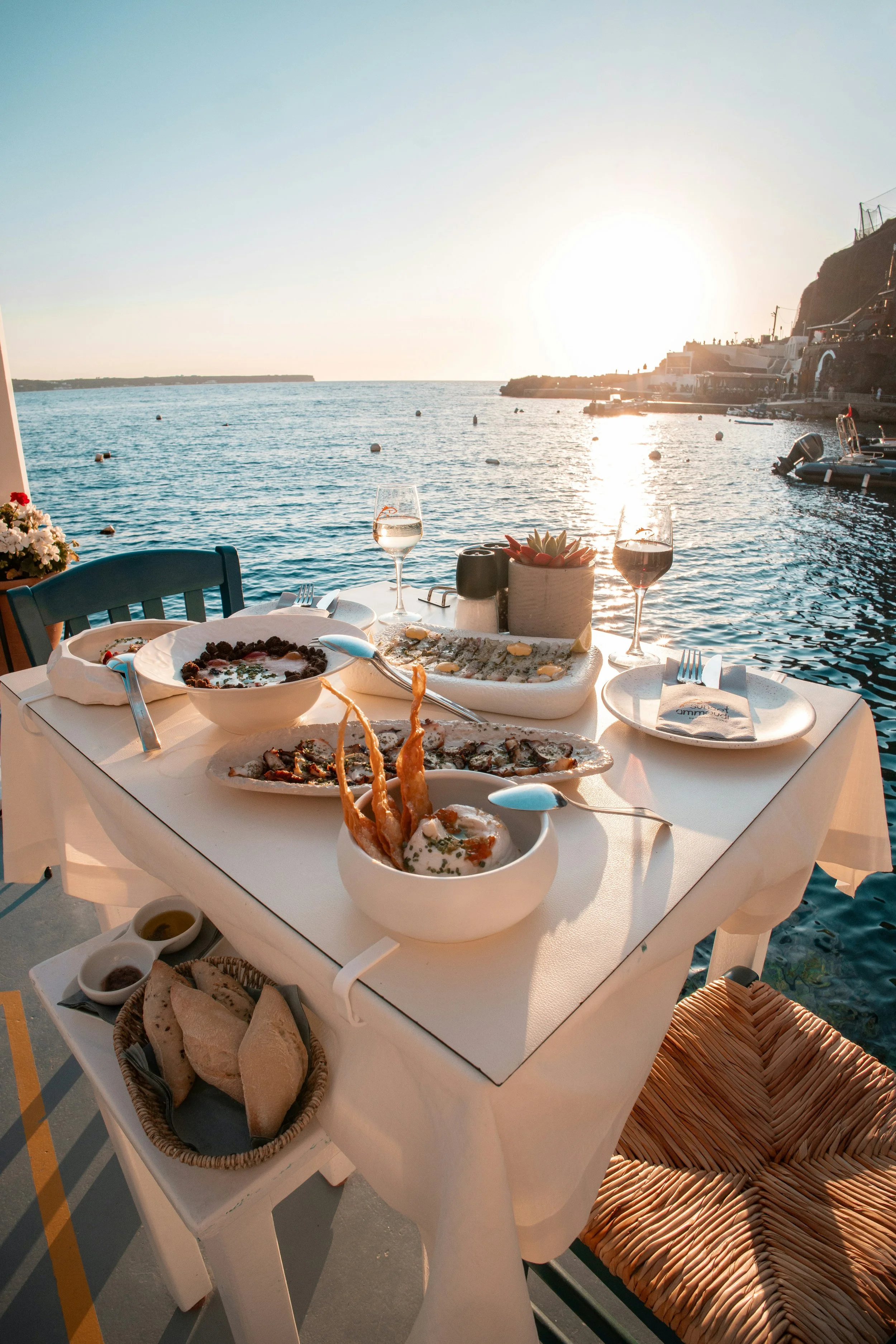A table set with Spanish dishes including paella, bread, and side sauces, situated outdoors near a harbor with boats and buildings, during sunset.