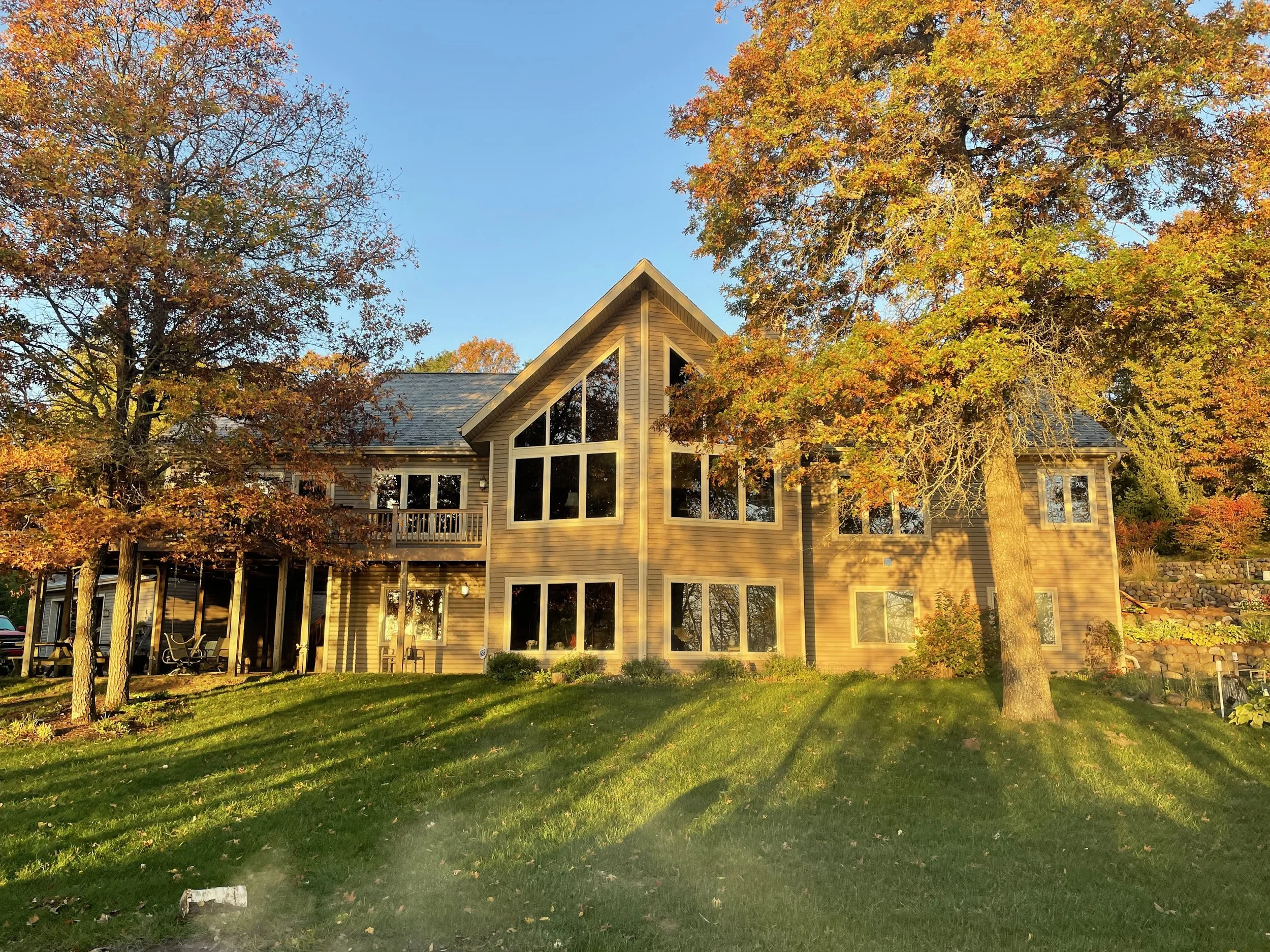 A house with large glass windows and a peaked roof is surrounded by trees with autumn-colored leaves. The house is illuminated by sunlight, and the lawn in front is green with shadows from the trees.