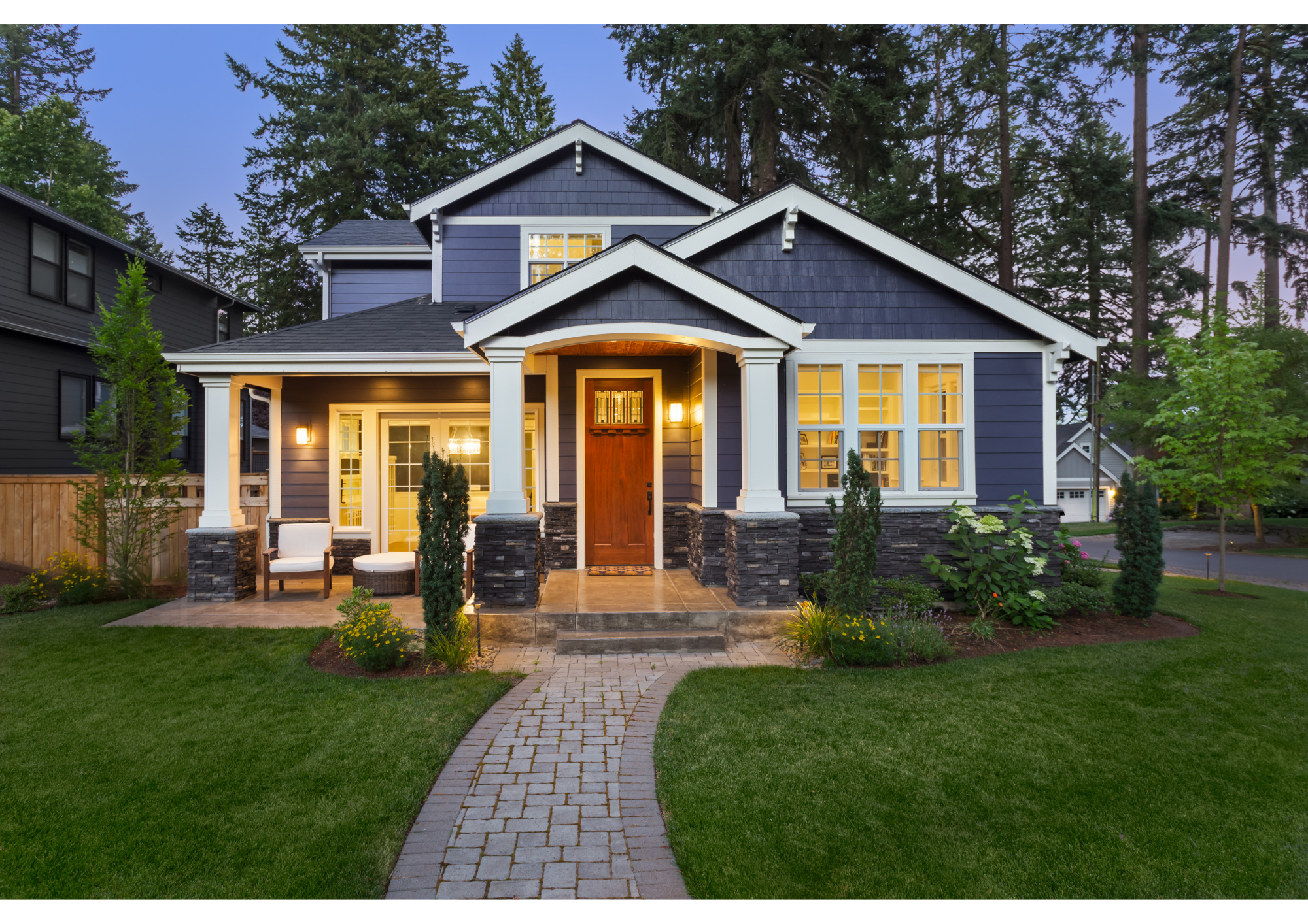 Front view of a modern house with a curved stone walkway leading to the front door, surrounded by well-maintained green grass, small trees, and decorative plants, with warm interior lighting visible through large windows and a porch area with seating.