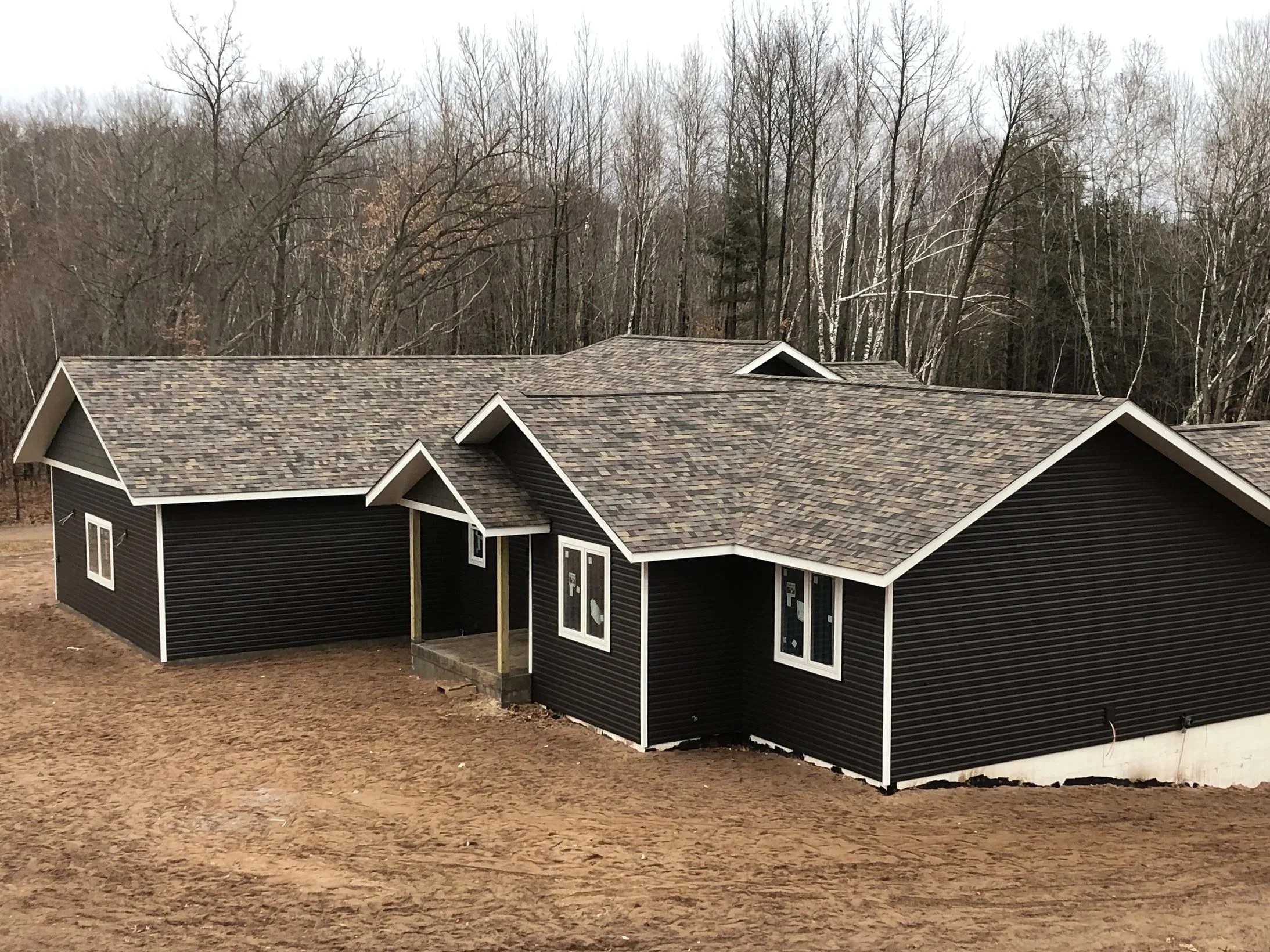 A house under construction with dark siding, multiple windows, and a multiple peaked roof, set against a background of leafless trees and an overcast sky.