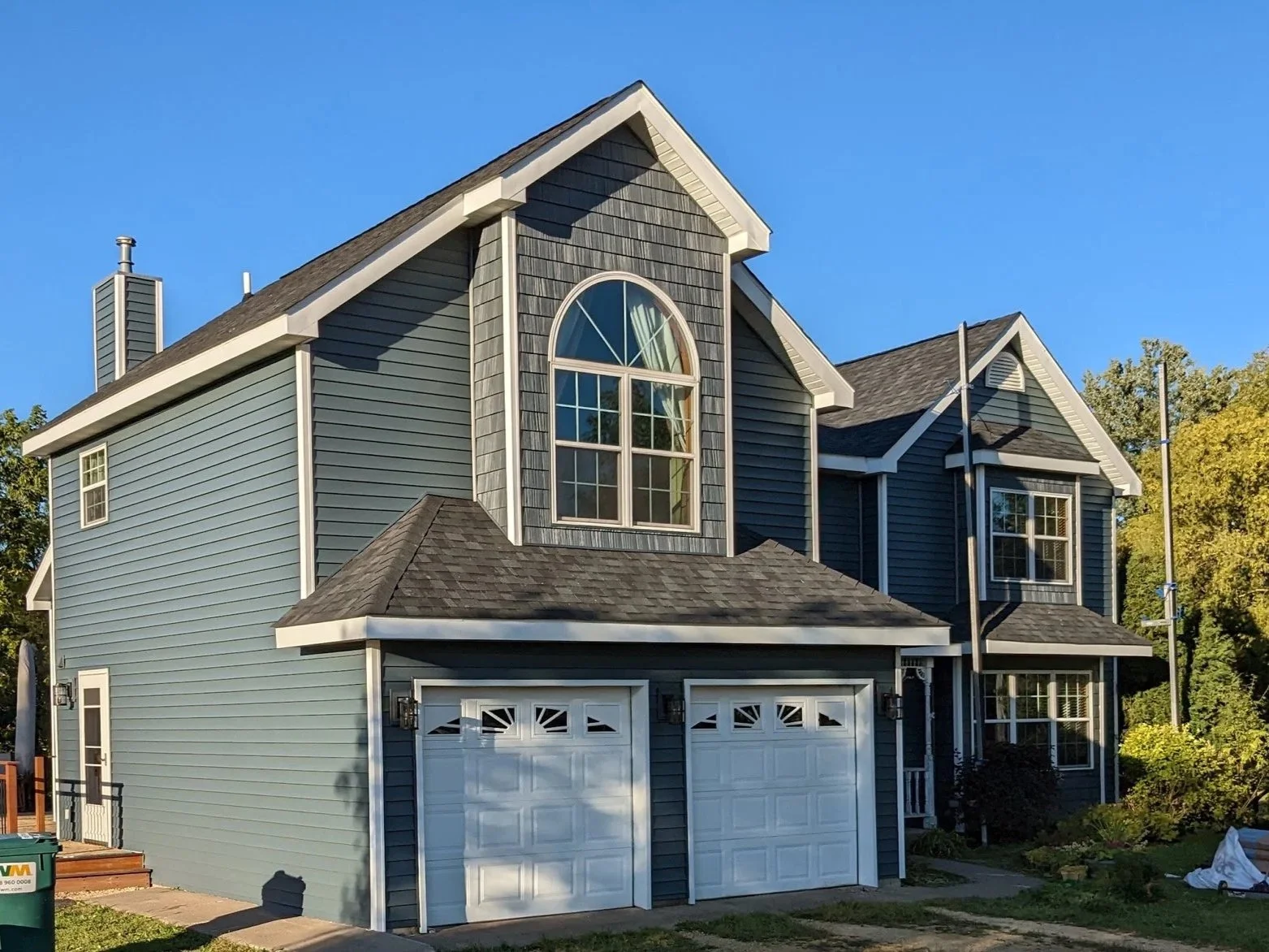 A two-story house with blue siding, a large arched window on the upper floor, and two white garage doors at the front. The house has a gable roof with dark shingles and is surrounded by trees and a clear blue sky.