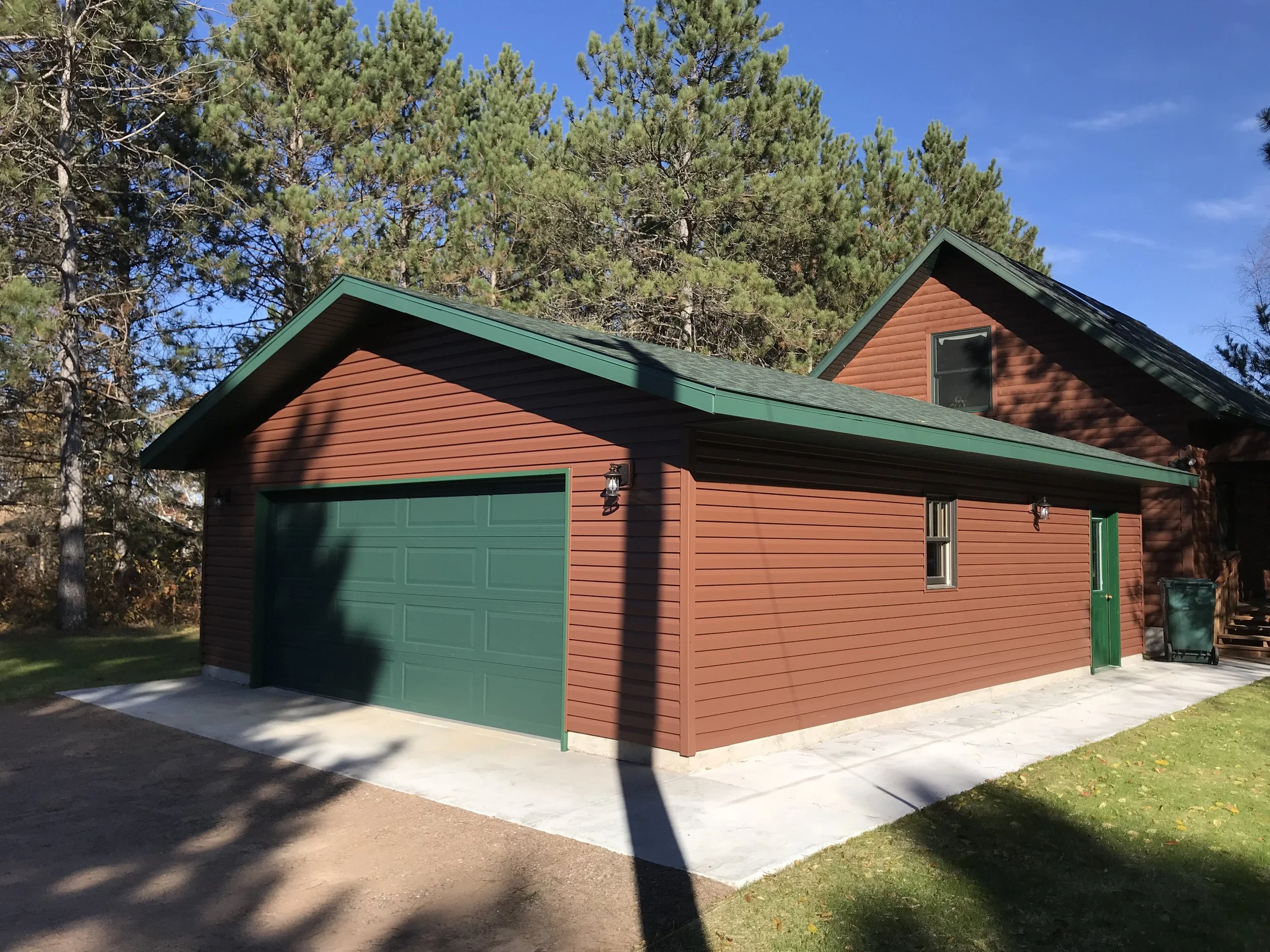 A red garage with green doors and trim, situated adjacent to a house with wood siding. The garage is on a concrete driveway with some shadows cast across it. In the background, there are tall pine trees and a clear blue sky.