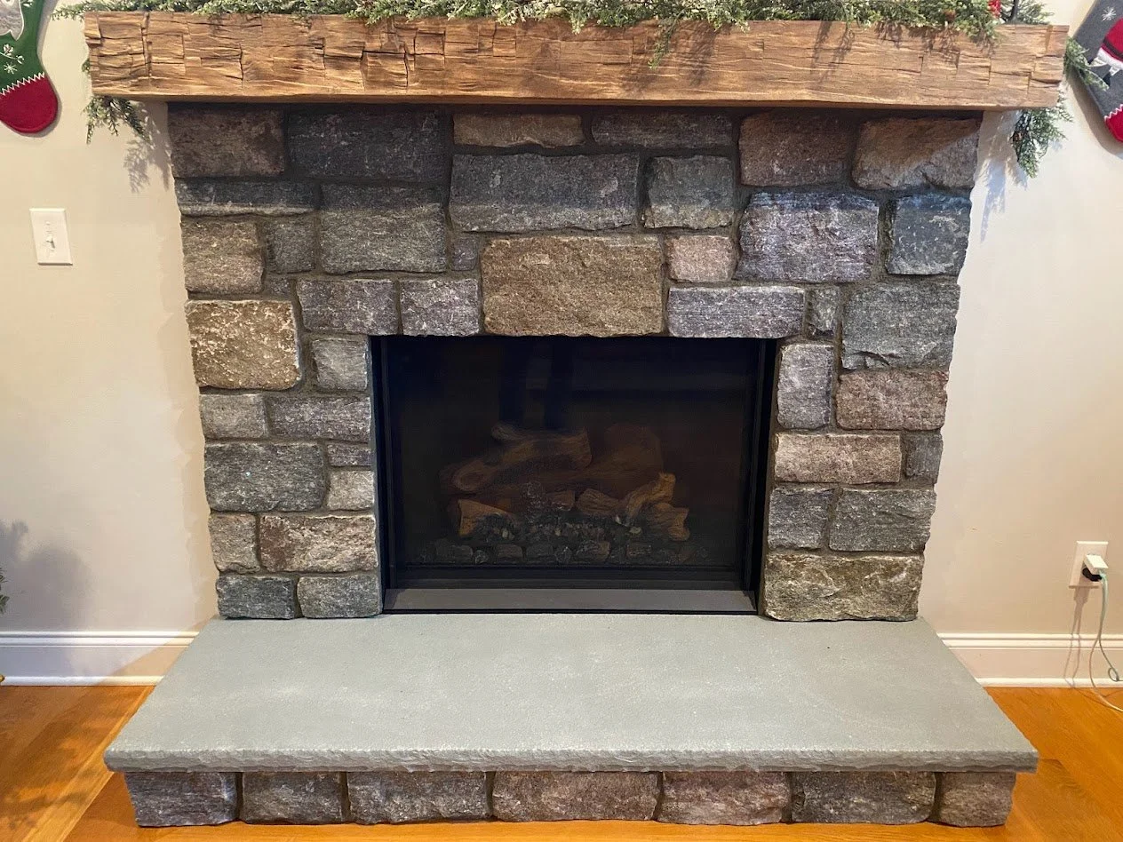A rustic fireplace made of large grey and brown stones with a concrete hearth and a wooden mantel decorated with greenery and small holiday decorations.
