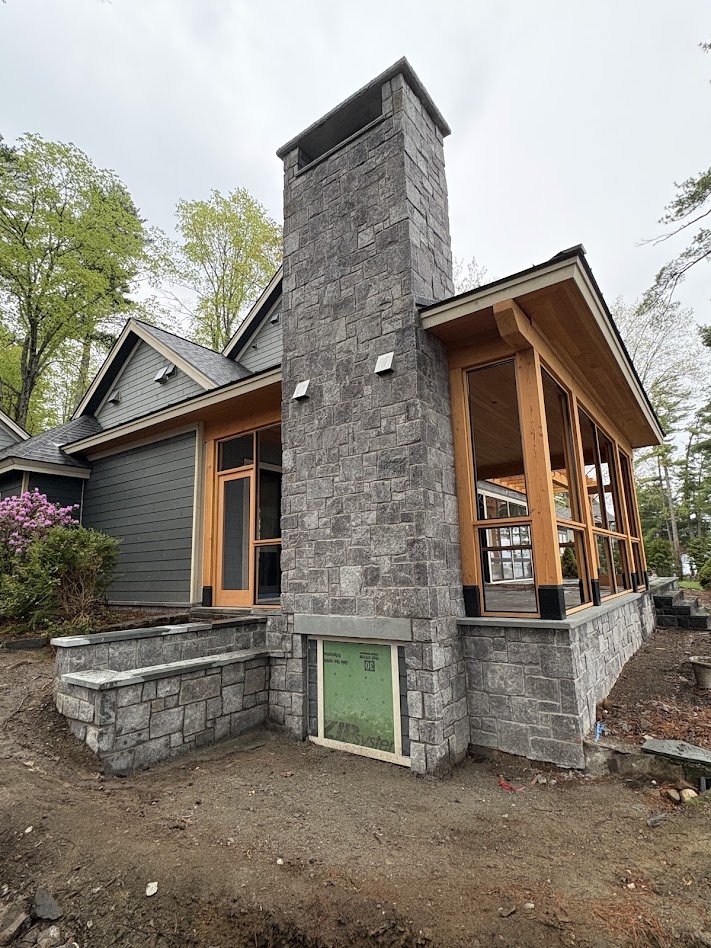 Exterior view of a house under construction featuring a large stone chimney, gray siding, wooden window frames, and a stone foundation.