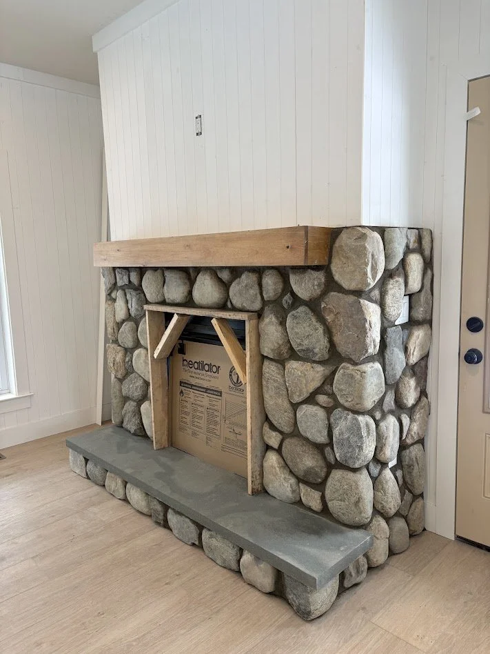 A stone fireplace with a newly built wooden mantel and a concrete hearth in a room with white paneled walls and light wood flooring.