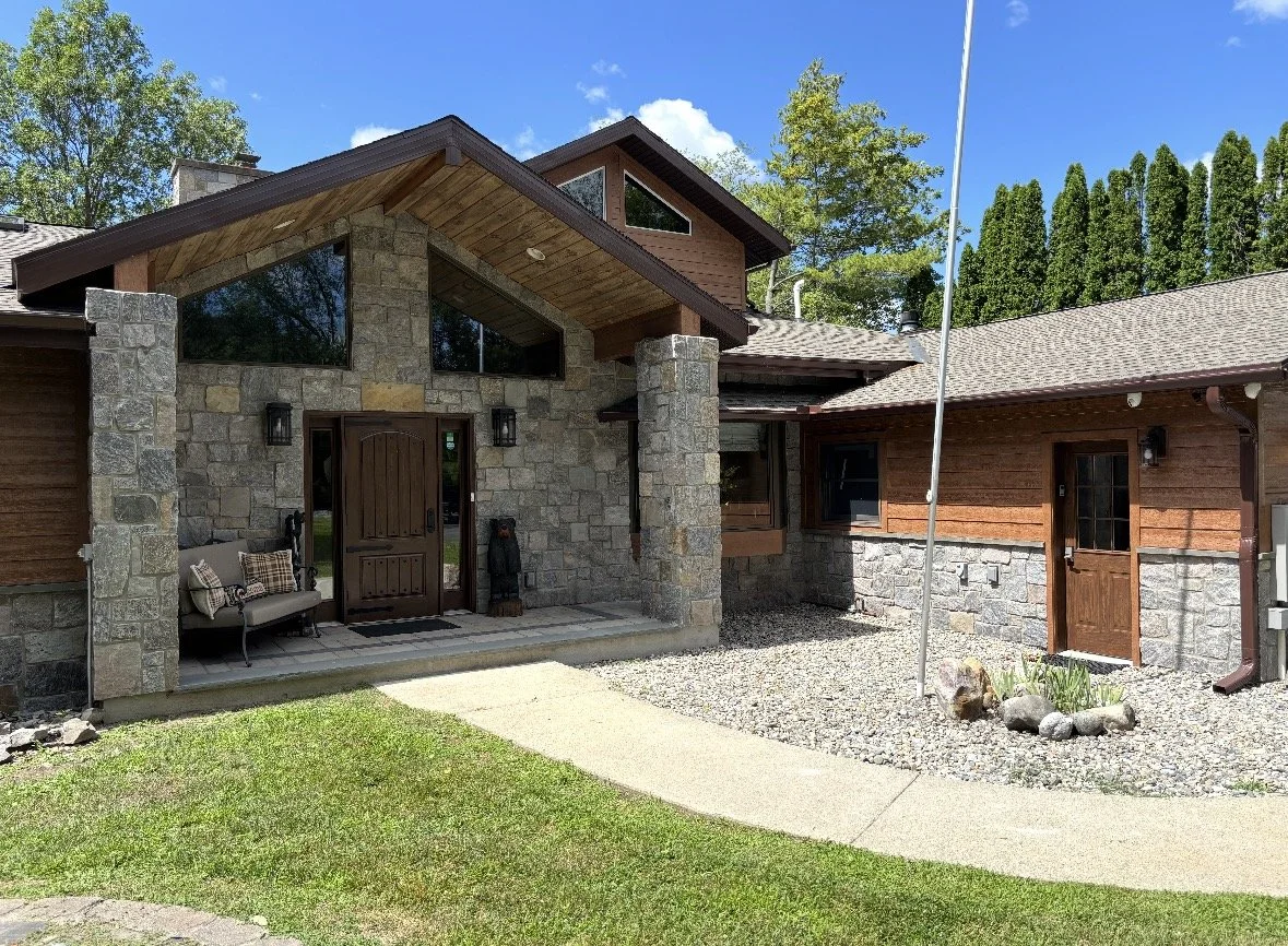 Exterior view of a house with stone and wood siding, a porch with seating, and a small garden bed, under a blue sky with some clouds.