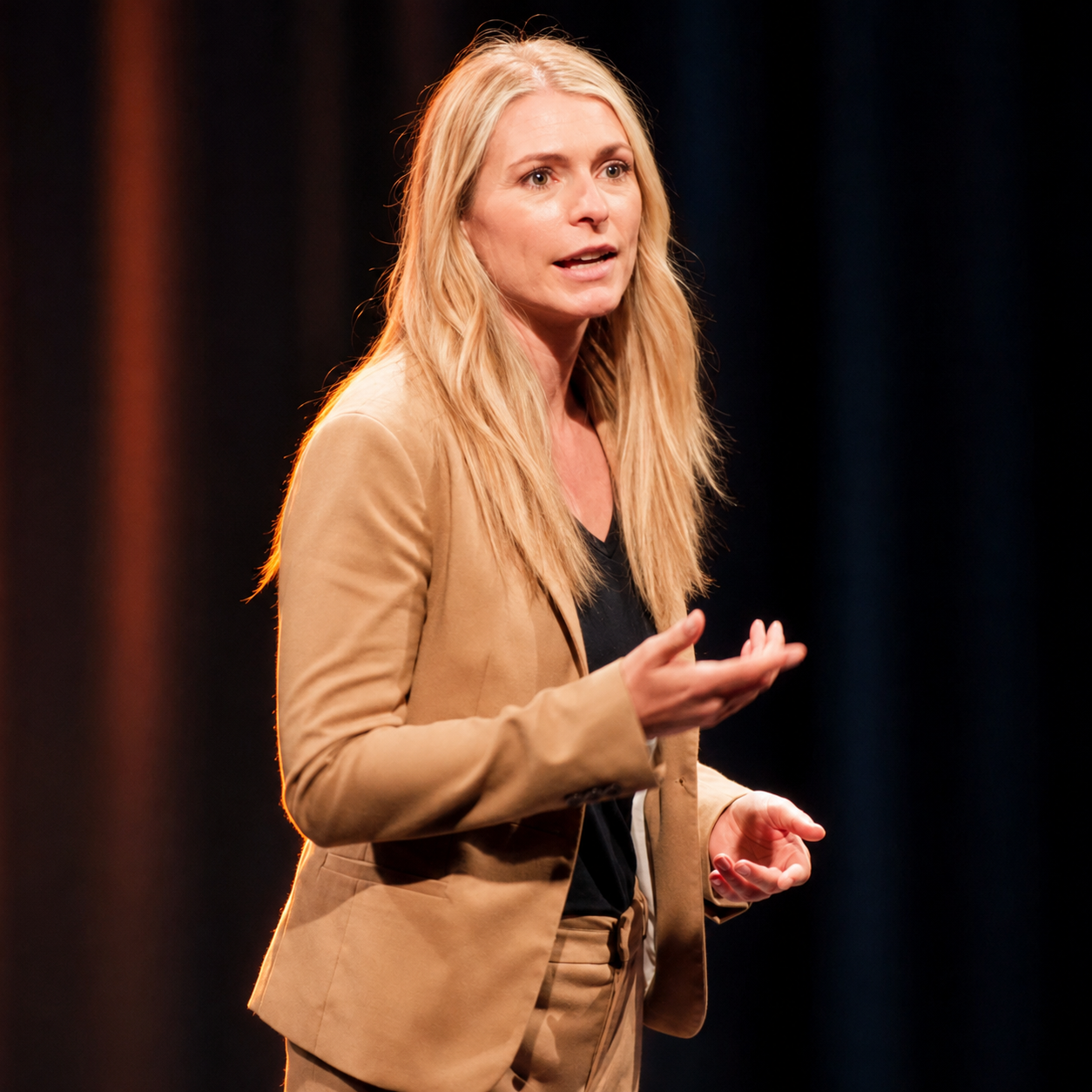 Sara Capra wearing a tan blazer and black top giving a speech on stage with dark curtains in the background.