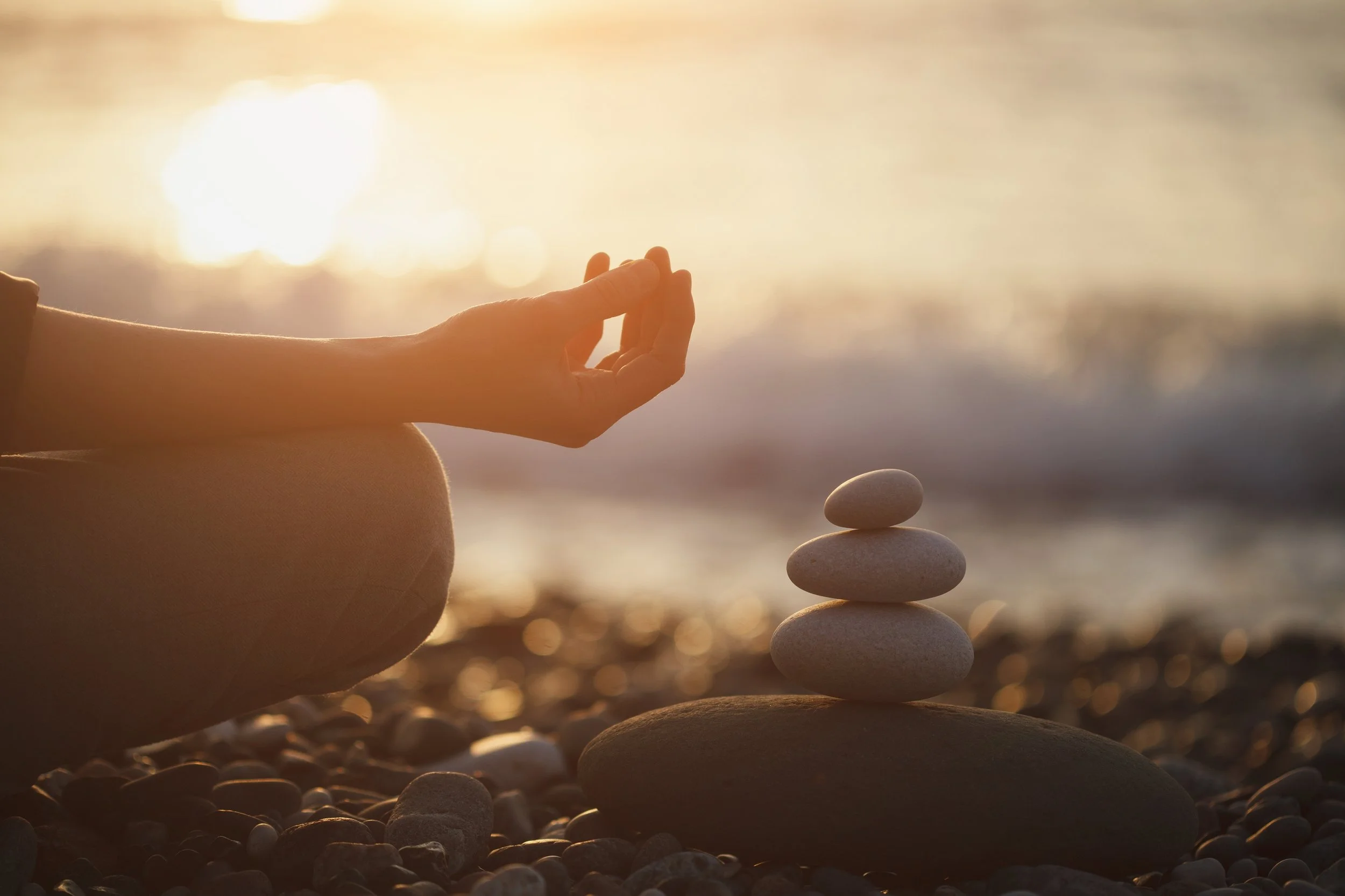 A person meditating on a beach at sunset, with a stack of balanced stones in the foreground.