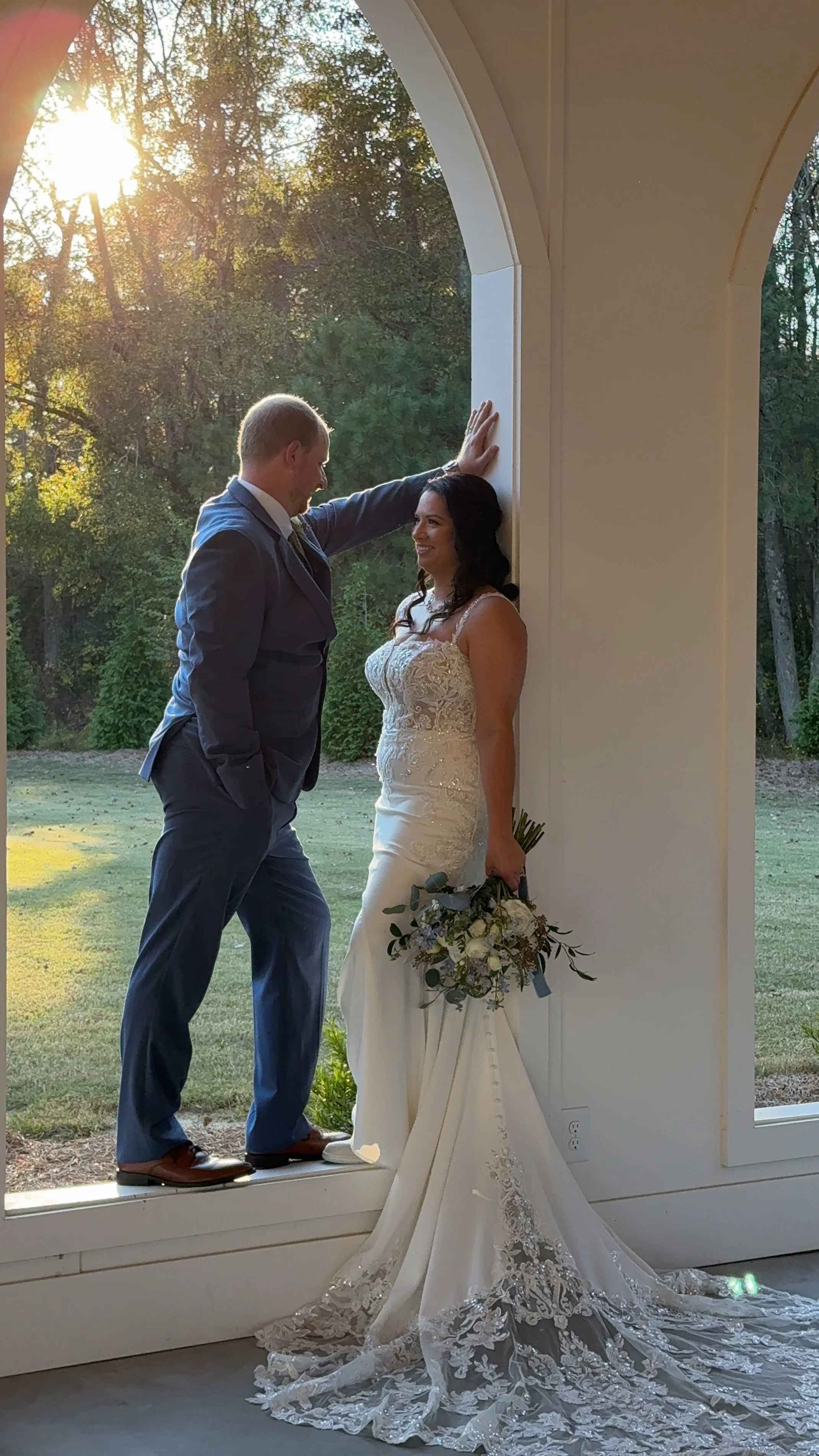 A bride and groom standing near a large window with sunlight shining through trees outside. The groom is leaning against the frame with one hand, smiling at the bride who holds a bouquet of flowers and wears a white lace wedding dress. The bride is smiling back at the groom.