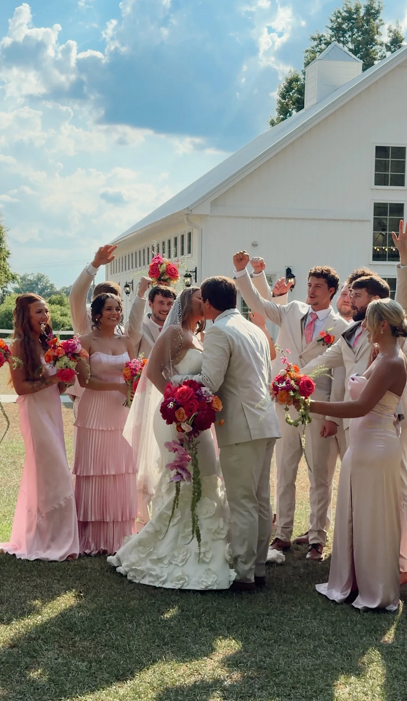 A wedding ceremony outdoors with a bride and groom kissing, surrounded by bridesmaids and groomsmen holding colorful bouquets and celebrating. A white building and blue sky with clouds are in the background.