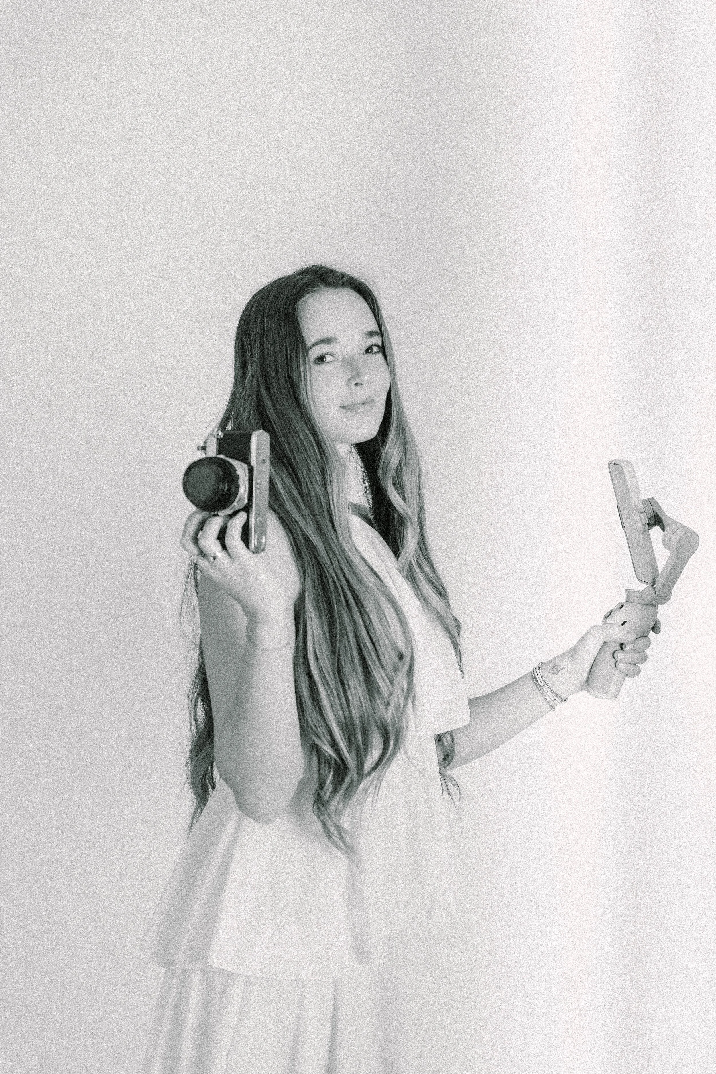 A woman with long wavy hair holding a vintage camera in her right hand while looking at the camera, wearing a white dress and jewelry, standing against a plain background.