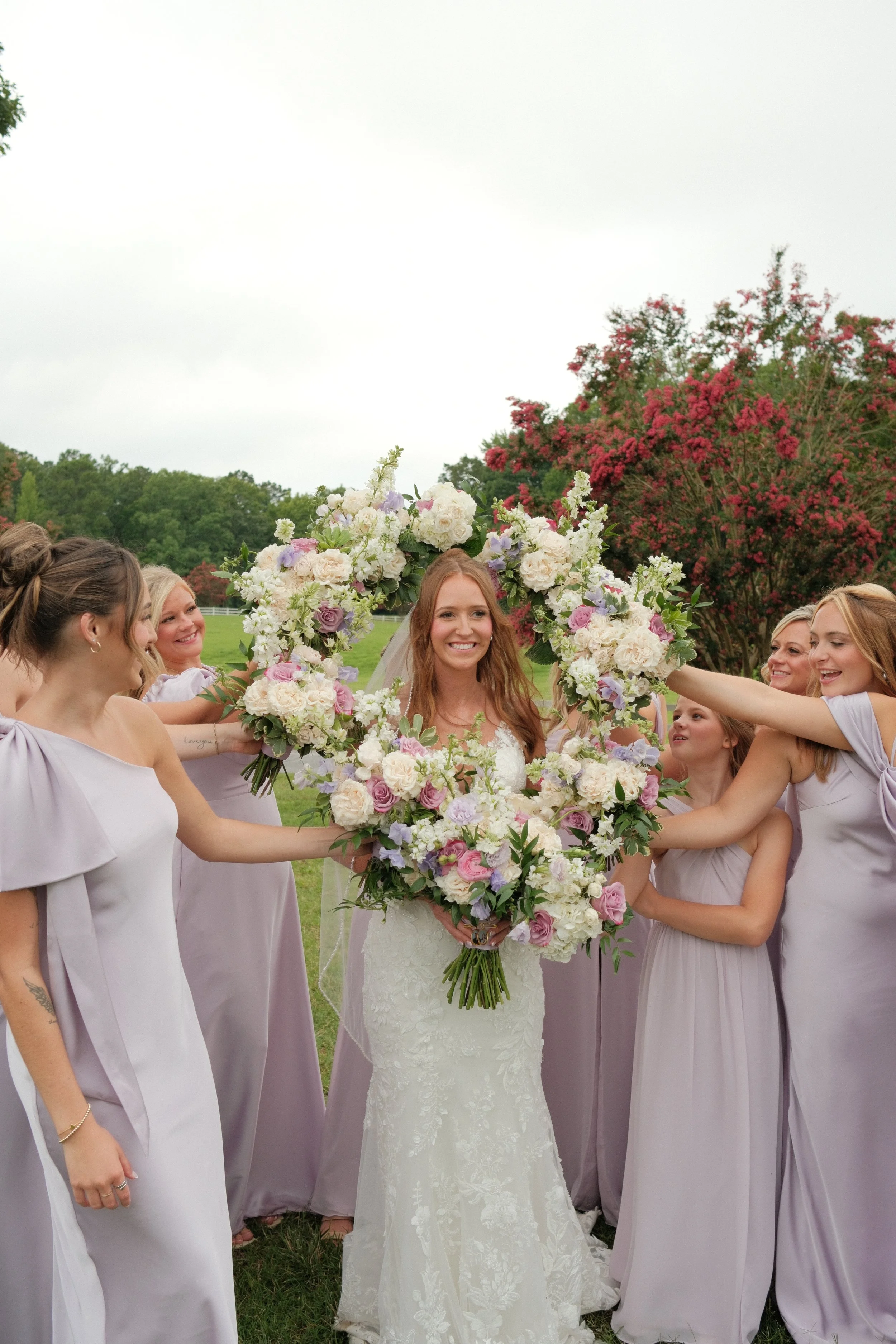 A bride surrounded by women in lavender bridesmaid dresses holding a large floral wedding bouquet outdoors on a lawn with trees and a flowering tree in the background.