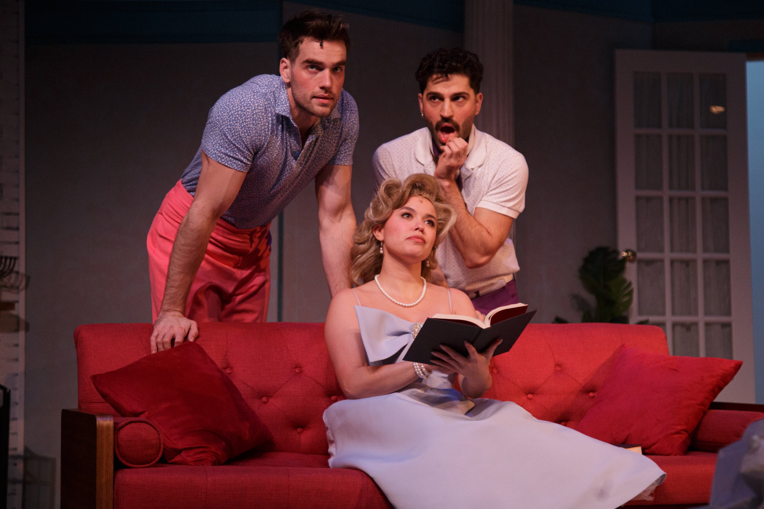 Three people, two men and a woman, are on a red couch, with the woman reading a book. The men are leaning over her, one with a curious expression and the other with a surprised or shocked expression. The scene looks like a performance or a staged scene indoors.