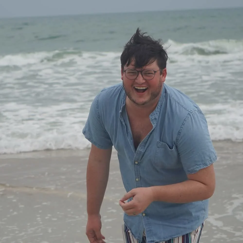 A man wearing glasses and a blue shirt is laughing on a beach with waves in the background.