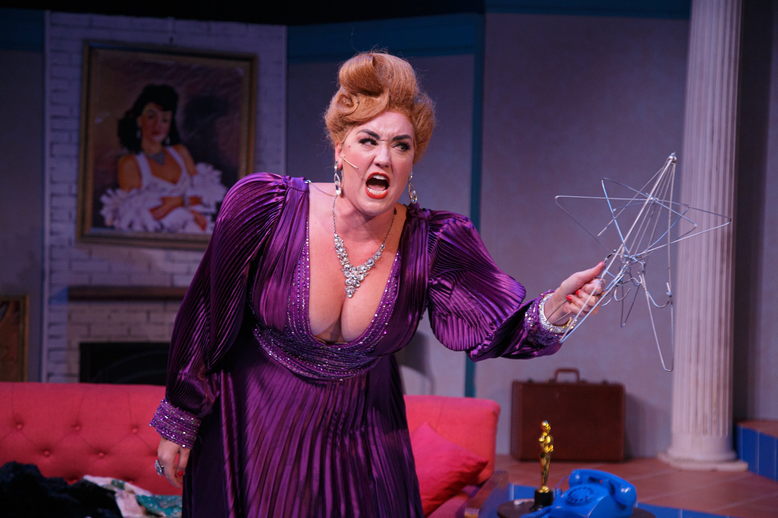 A woman with red hair styled in an elaborate updo, wearing a purple dress with a plunging neckline and jewelry, appears to be angry or yelling while holding a wire sculpture. She is inside a room with a framed portrait of a woman wearing a white dress on the wall behind her, and a red couch with some clothes and objects on it in the background.