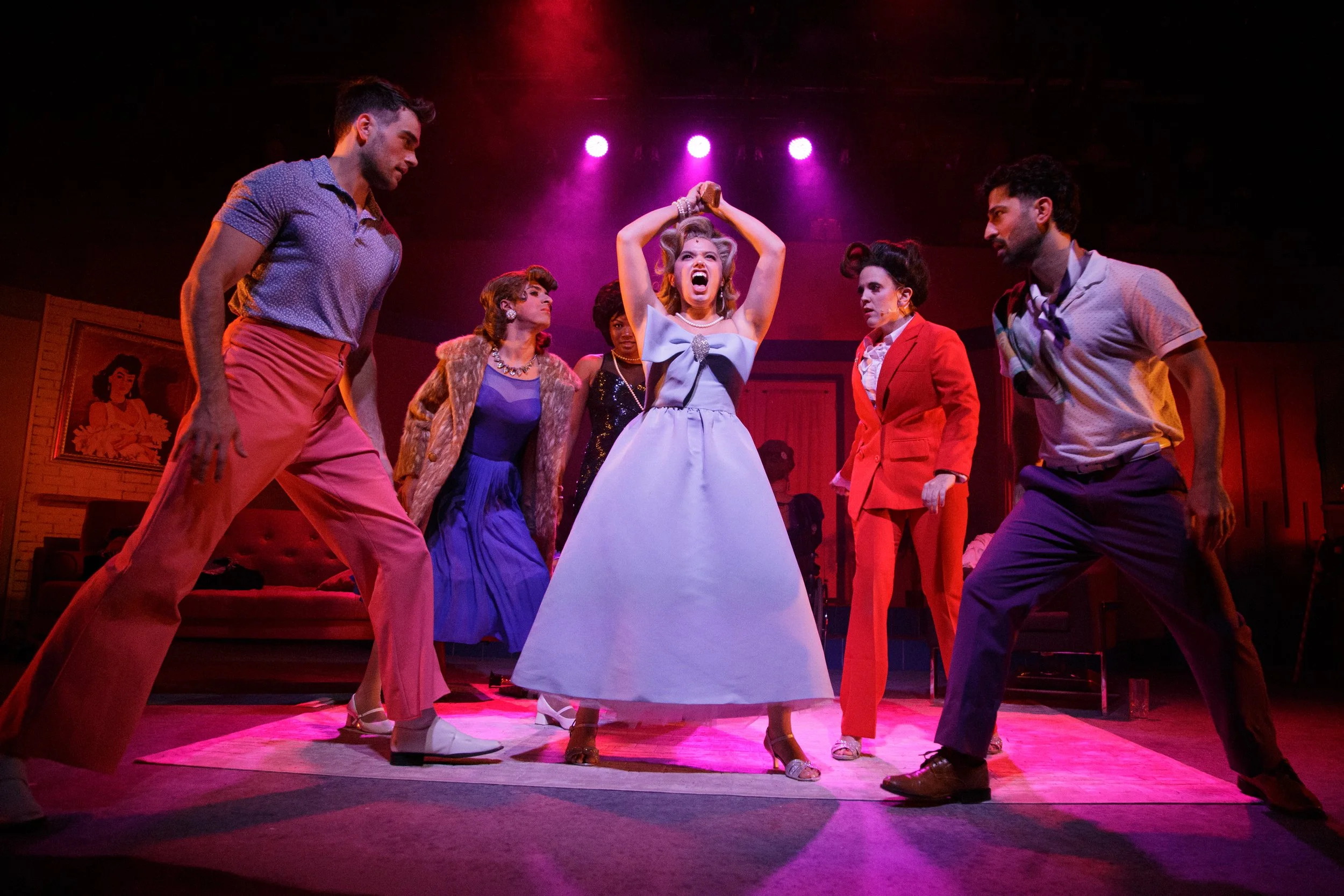 The image shows a theatrical scene with six actors on stage, dressed in vintage clothing from the 1950s or 1960s. The central female character appears angry or distressed, with her arms raised above her head. The surrounding actors are intently focused on her, with some appearing stern or confrontational. The stage is lit with pink and purple lights, and there is a painting of a woman in the background.