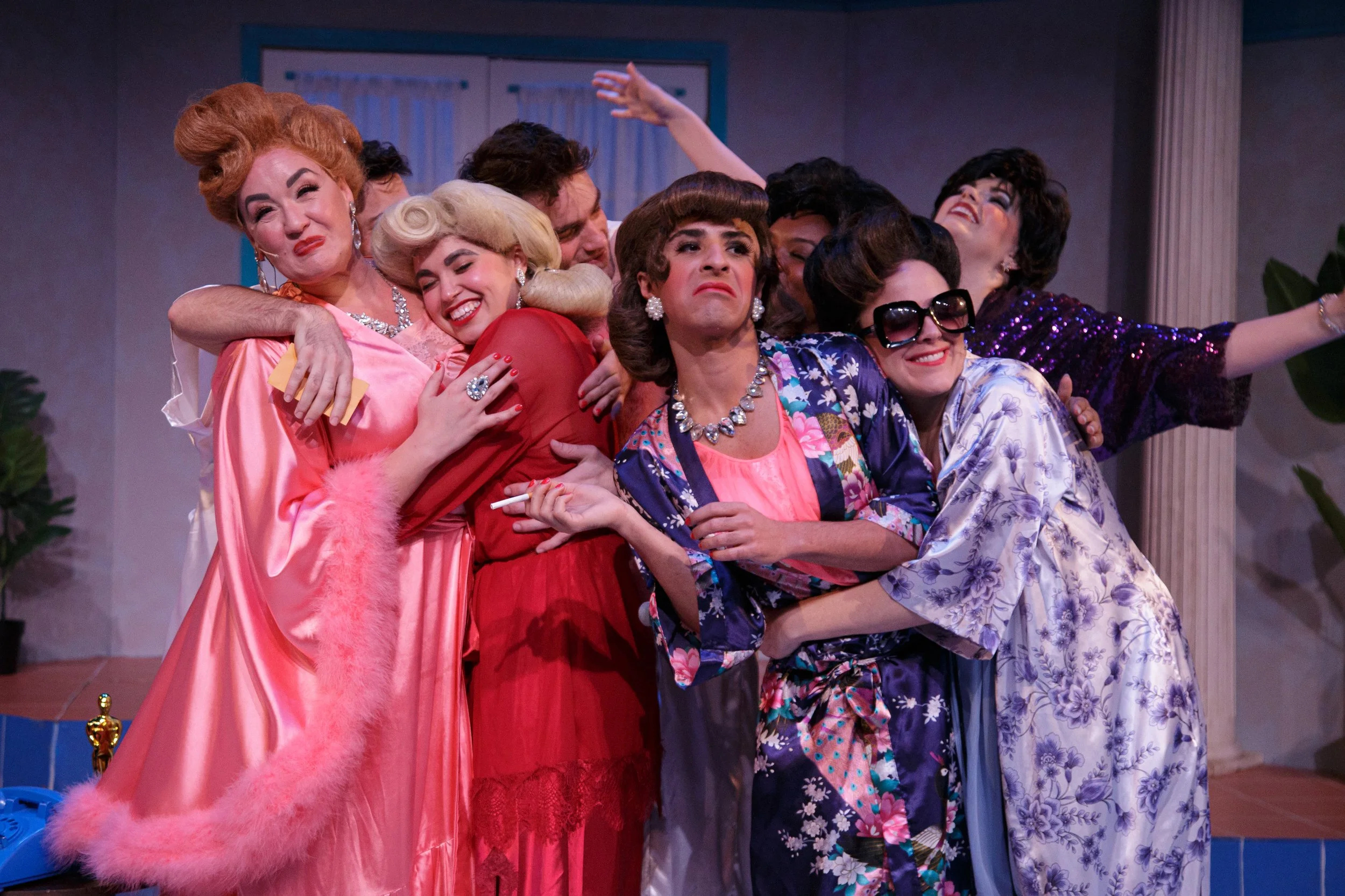 Group of women in colorful dresses and patterns, some with jewelry and sunglasses, hugging and smiling on a stage with a blue background and curtains.
