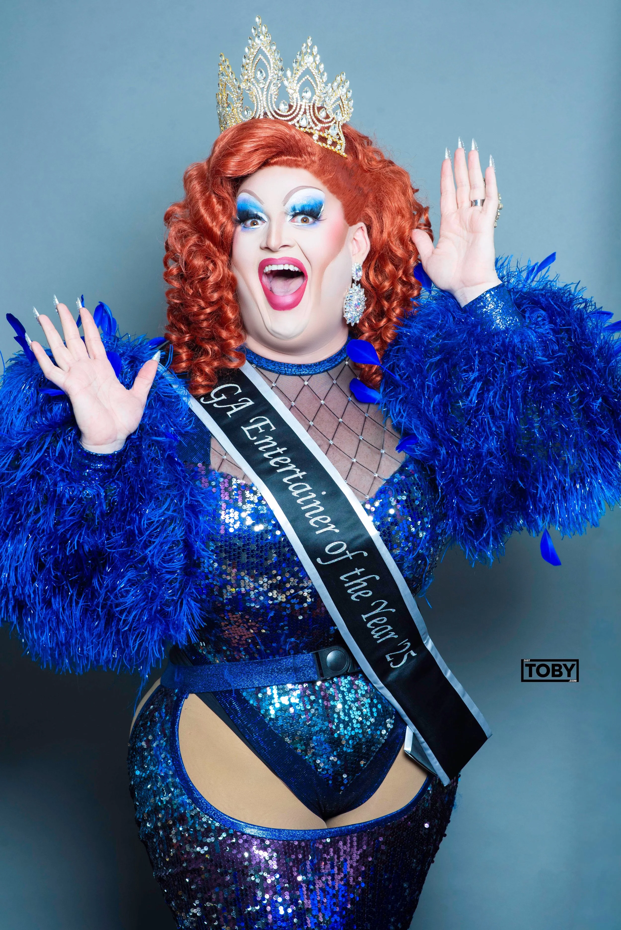 A person dressed in a glamorous, sequined blue dress with cutouts, wearing a sash that reads 'Gae Entertainment of the Year 25', a large crown, and dramatic makeup. The person has red curly hair and is posing with an expressive face and hand gestures against a plain background.