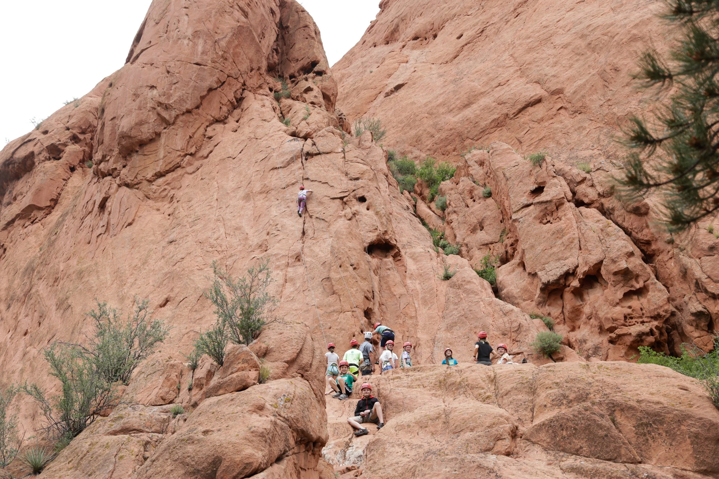 A group of people in helmets sitting on rocks at the base of a tall red sandstone cliff, some climbing or resting while others wait or observe.