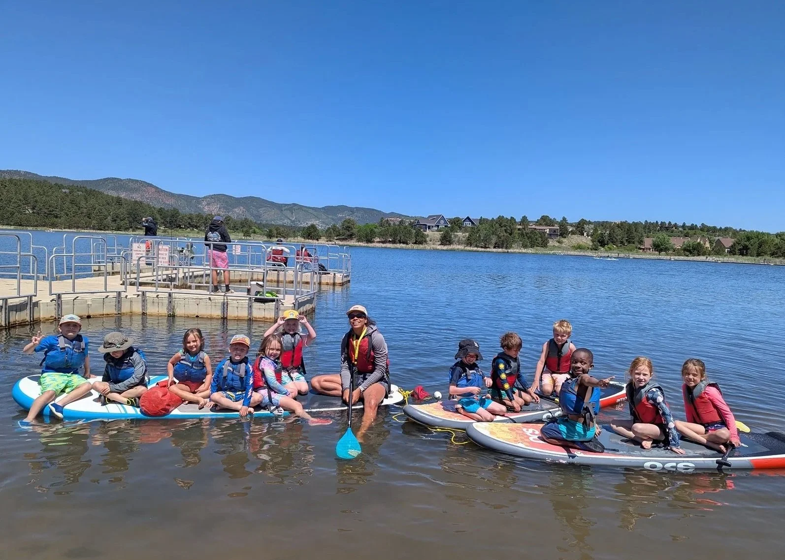 Group of children and an adult sitting on paddleboards in a lake with a dock and boats in the background on a sunny day.