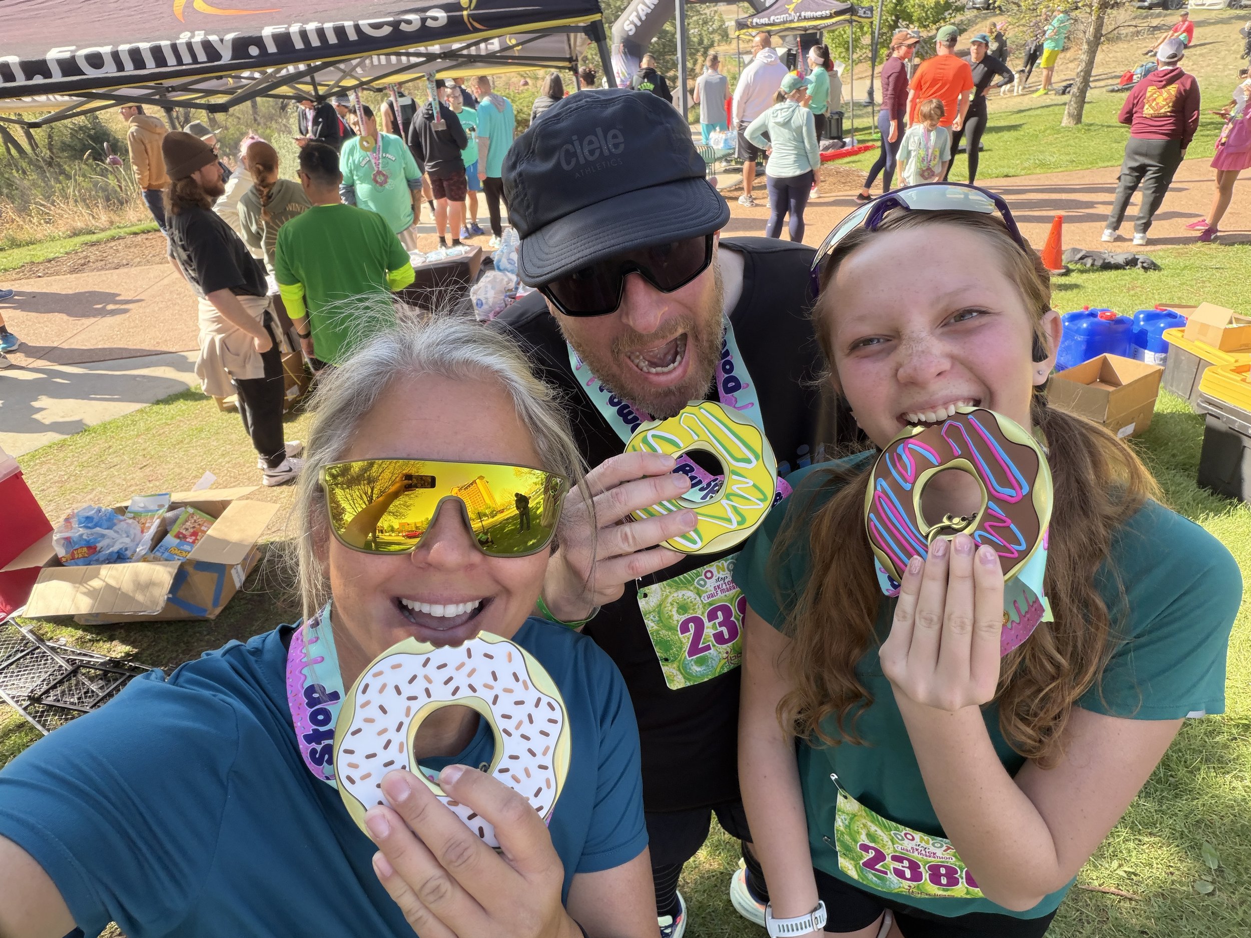 A group of people gathered outdoors at a fun run event, holding decorative donut-shaped medals, with some wearing race bibs. There are tents and many participants in the background, some are wearing colorful athletic clothing.