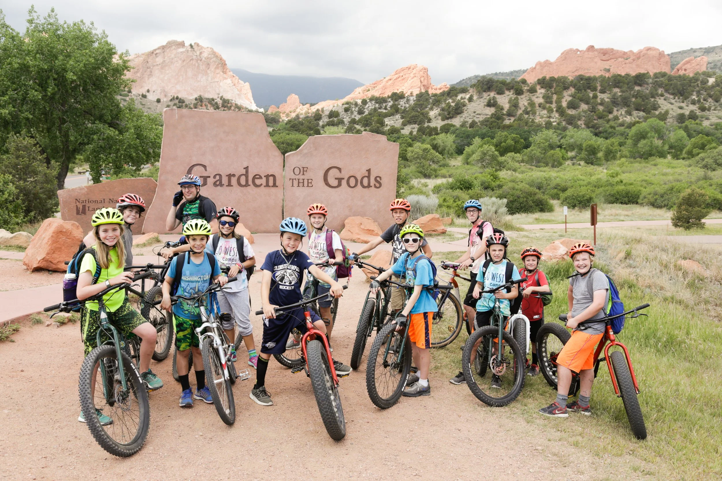 A group of children and their adult cycling companions posing with their bikes in front of the Garden of the Gods entrance sign in Colorado, with red rock formations and green trees in the background.