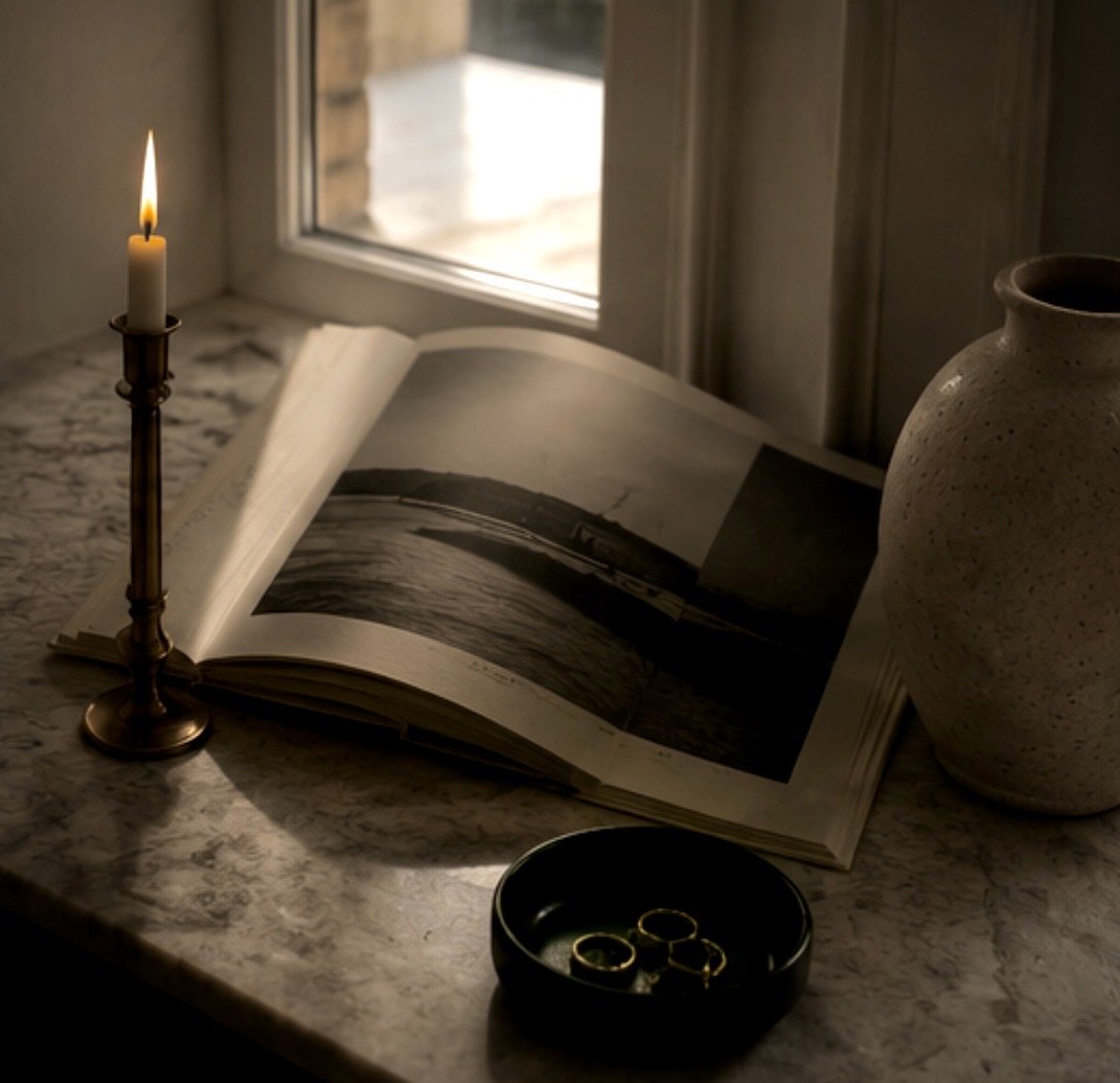 A lit candle, an open photo book, a ceramic vase, and three gold rings in a black dish on a marble windowsill.