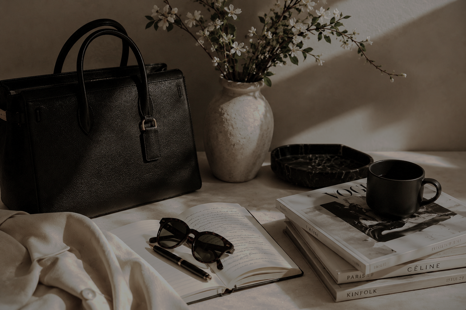 A black leather handbag, a vase with white flowers, a black marble tray, a stack of fashion magazines, a black coffee mug, an open notebook with sunglasses and a pen, and a cream-colored fabric are arranged on a light-colored table.