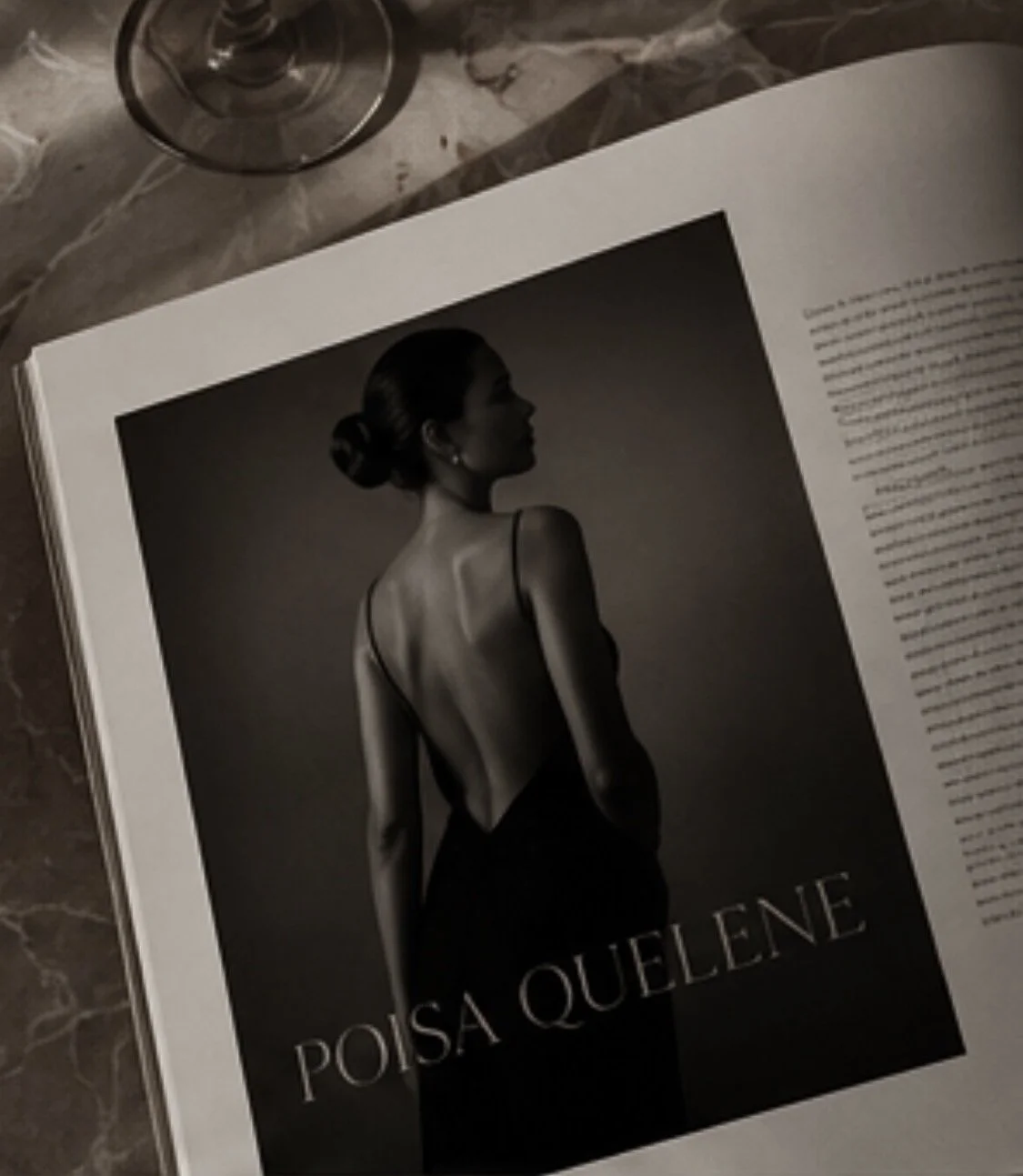 Open magazine featuring a black and white profile photo of a woman with her hair in a bun, wearing a backless dress, on a marble table near an empty wine glass.